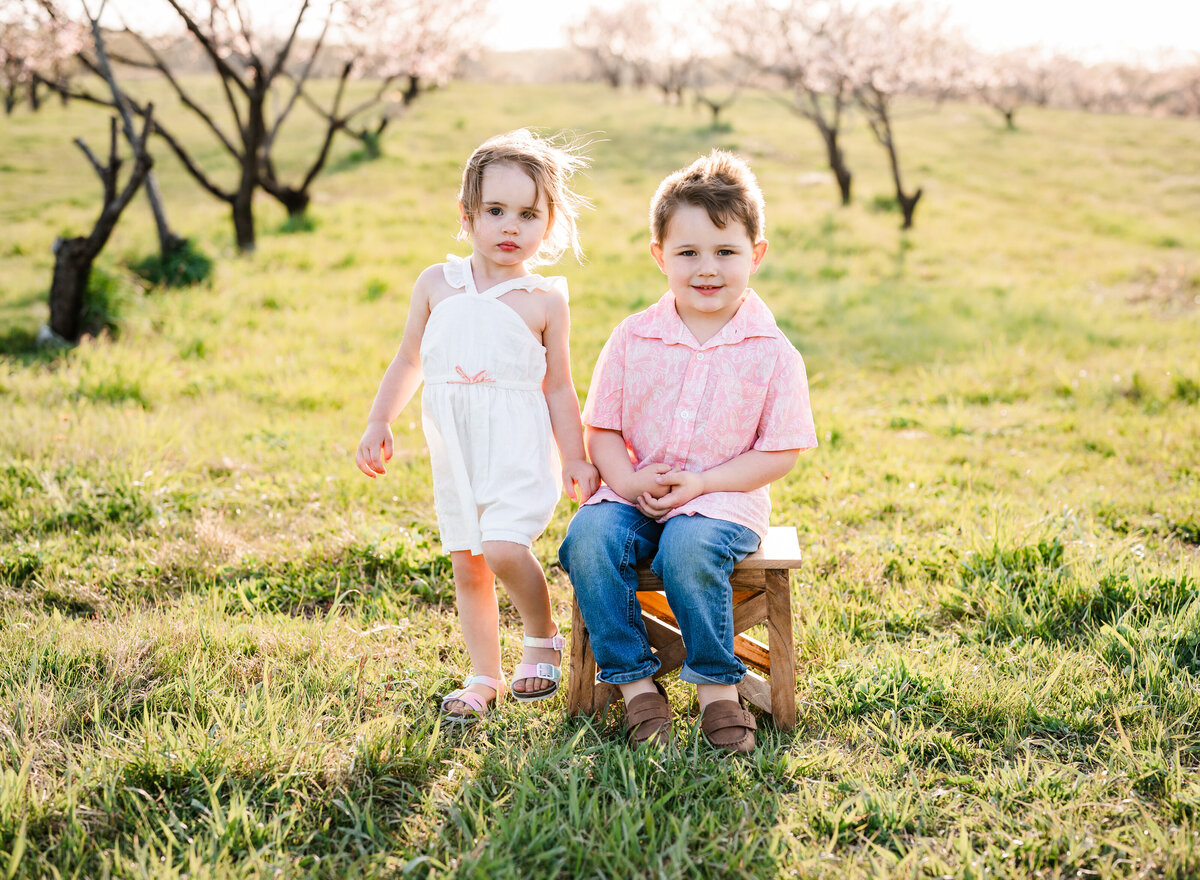 A boy and girl sitting on a small wooden bench in a grassy field during golden hour