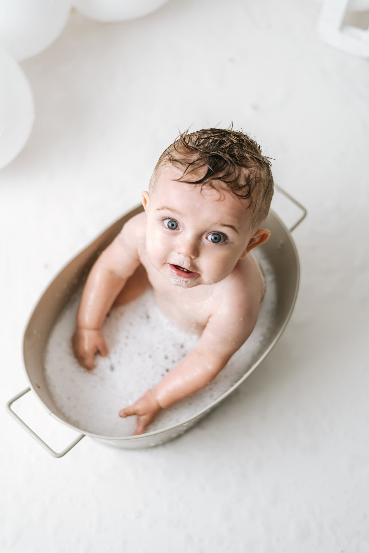 Baby boy sitting in a bath tub with bubbles looking up at a cake smash photoshoot in Billngshurst