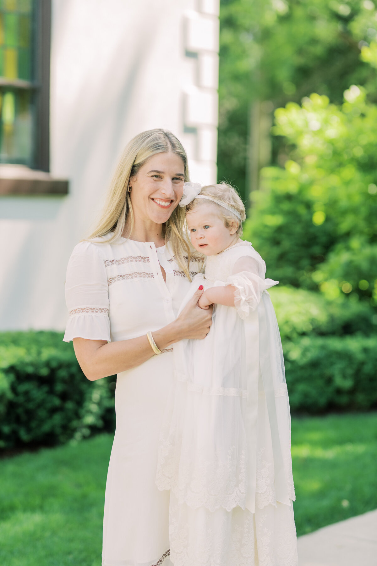 Mother in ivory dress holding her baby post-baptism, surrounded by garden hues and summer serenity.
