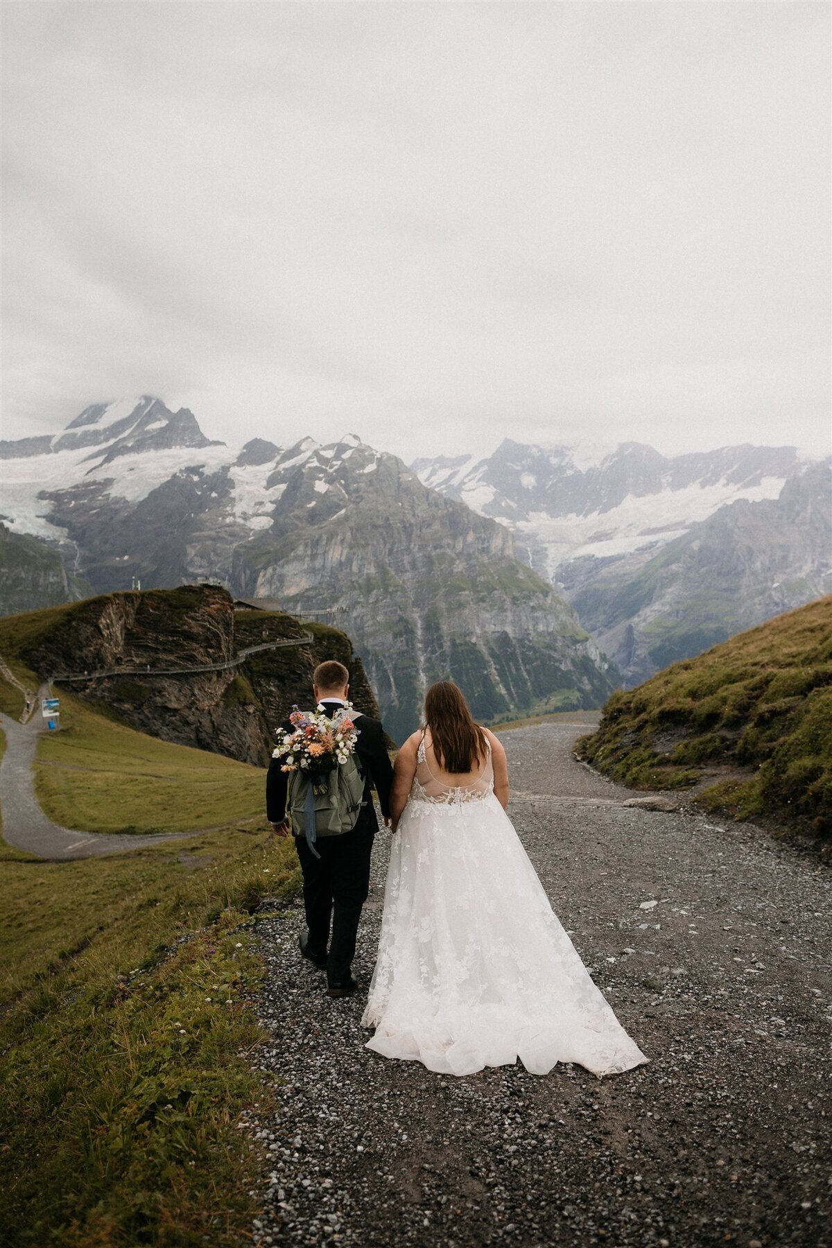 Bride & groom with bouquet in his back pack walking  and holding hands along hiking trail in the Swiss Alps!