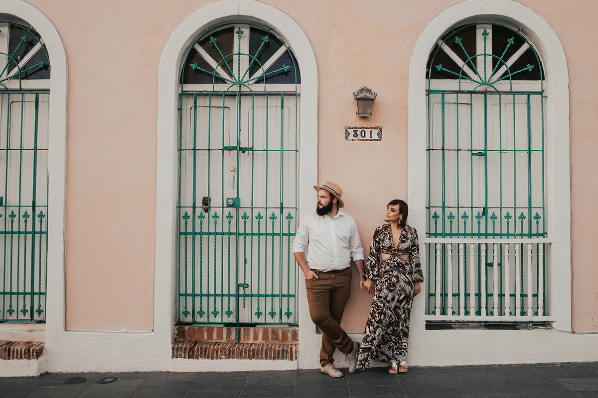 A couple holding hands and leaning against a pink building 