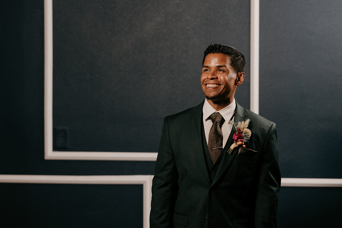The groom smiles broadly, looking off to the side in his dark green suit and floral boutonniere, standing against a sophisticated dark wall with white trim.