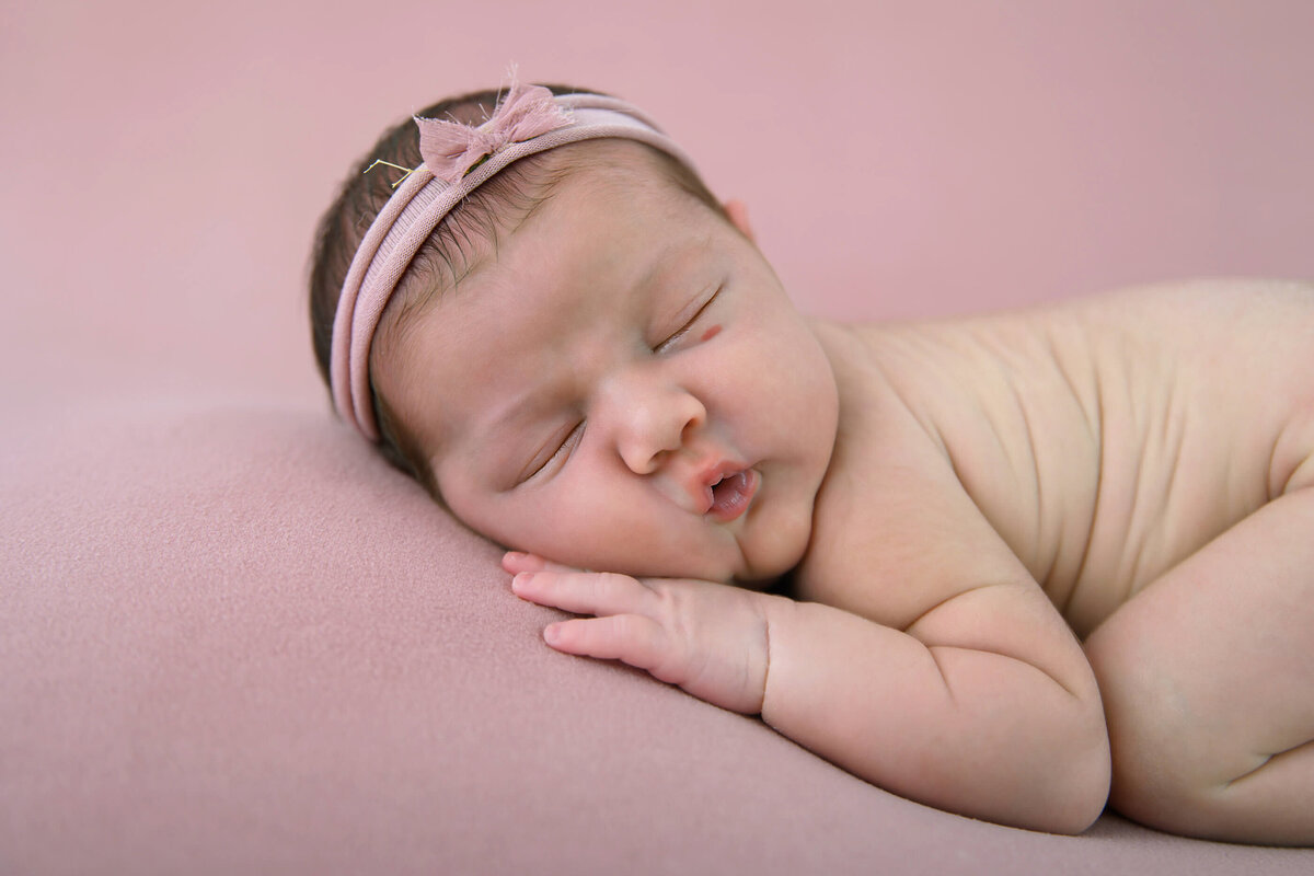 close up of newborn girl wearing a pink headband on a pink background for her newborn photos in Burlington