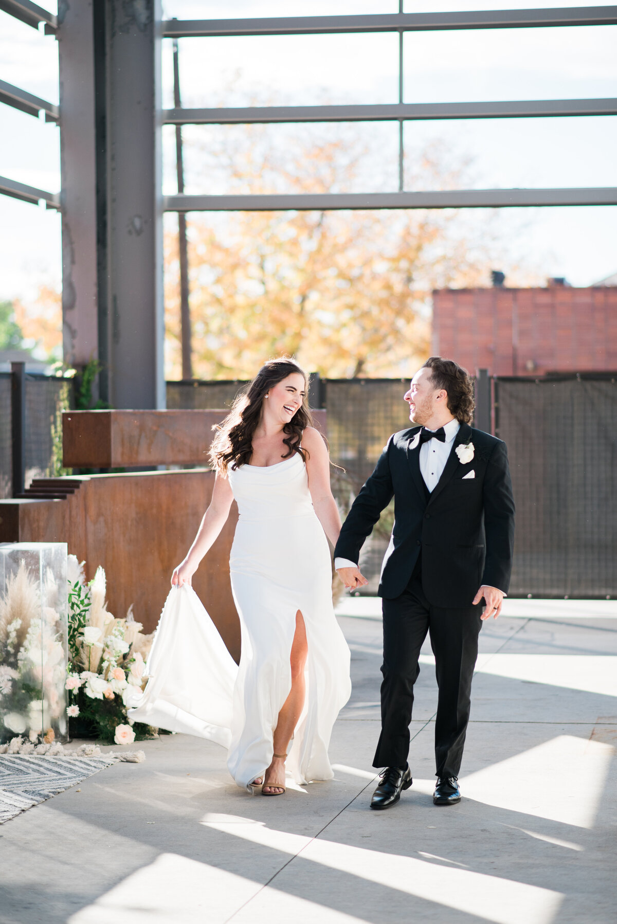 denver wedding photographer captures elegant wedding images of bride and groom holdin ghands as they walk through their urban colorado wedding venue with bride in a satin gown and groom in a tux