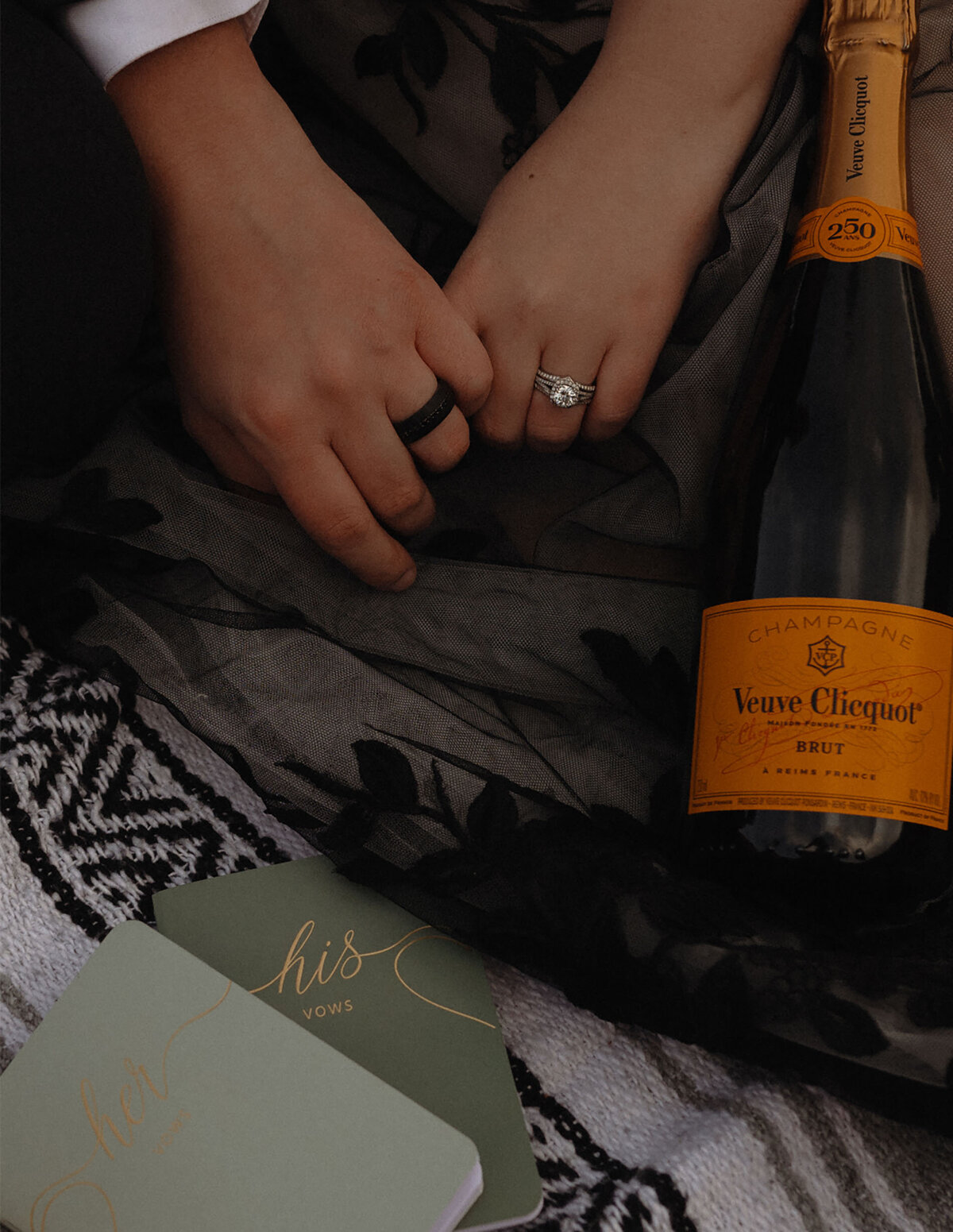 bride and groom hold hands and show off their wedding rings with a bottle of champagne sitting close by