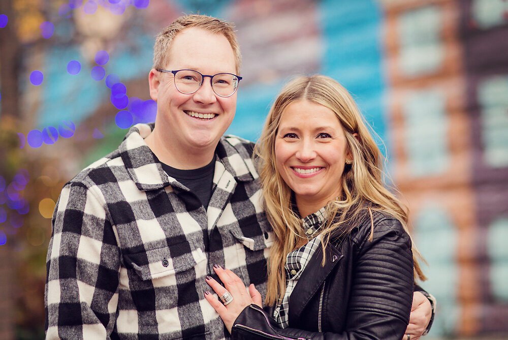 A couple is smiling in front of a colorful wall with Christmas lights in the background.