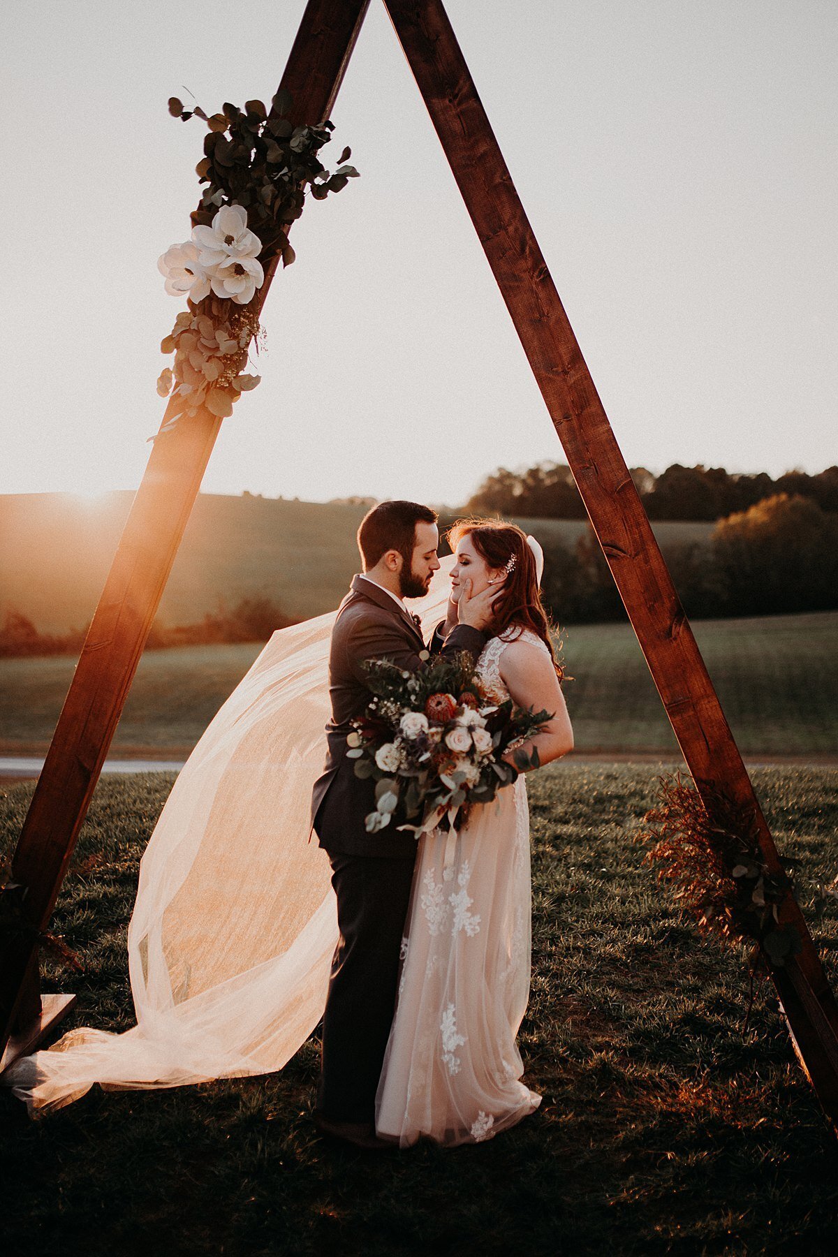 Standing under a triangular dark wood arbor decorated with silver dollar eucalyptus and magnolia blossoms, the bride and groom kiss at sunset. The bride is wearing a sheer blush wedding dress and is holding a large bouquet of red peonies, white roses, white anemonies, burgundy dahlia and eucalyptus.
