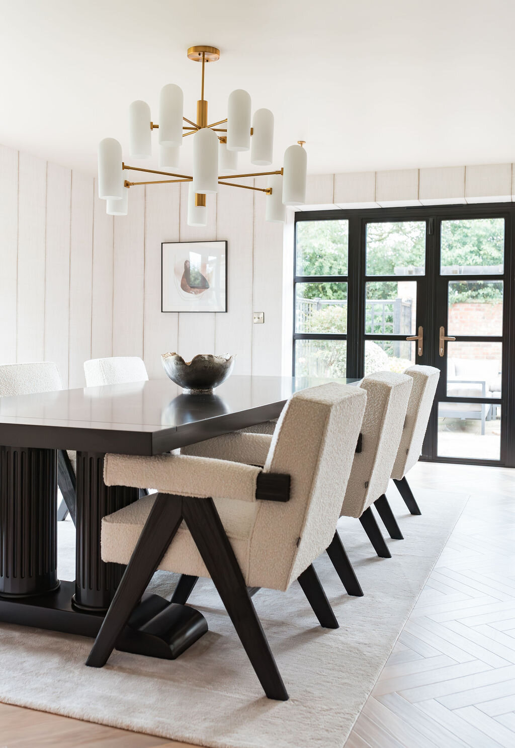 A modern dining room features a sleek black table surrounded by white upholstered chairs with black legs. A large white area rug covers the herringbone-patterned wooden floor. There is a gold chandelier and abundant natural light flooding through large windows.