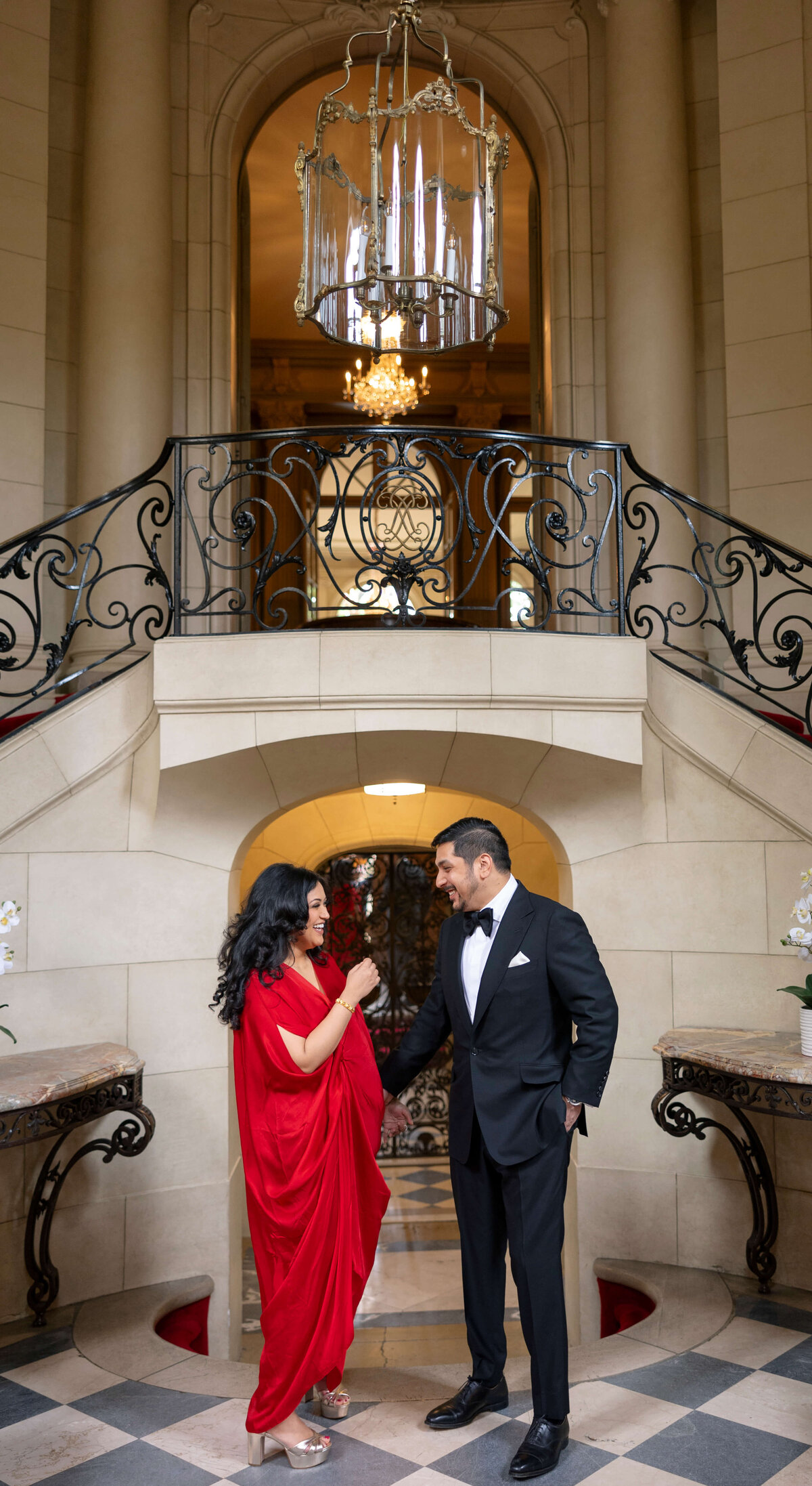 A woman in a red dress and a man in a black tuxedo stand at the base of a grand staircase with ornate railings. They are smiling at each other. A large chandelier hangs above them, and elegant decor completes the setting.