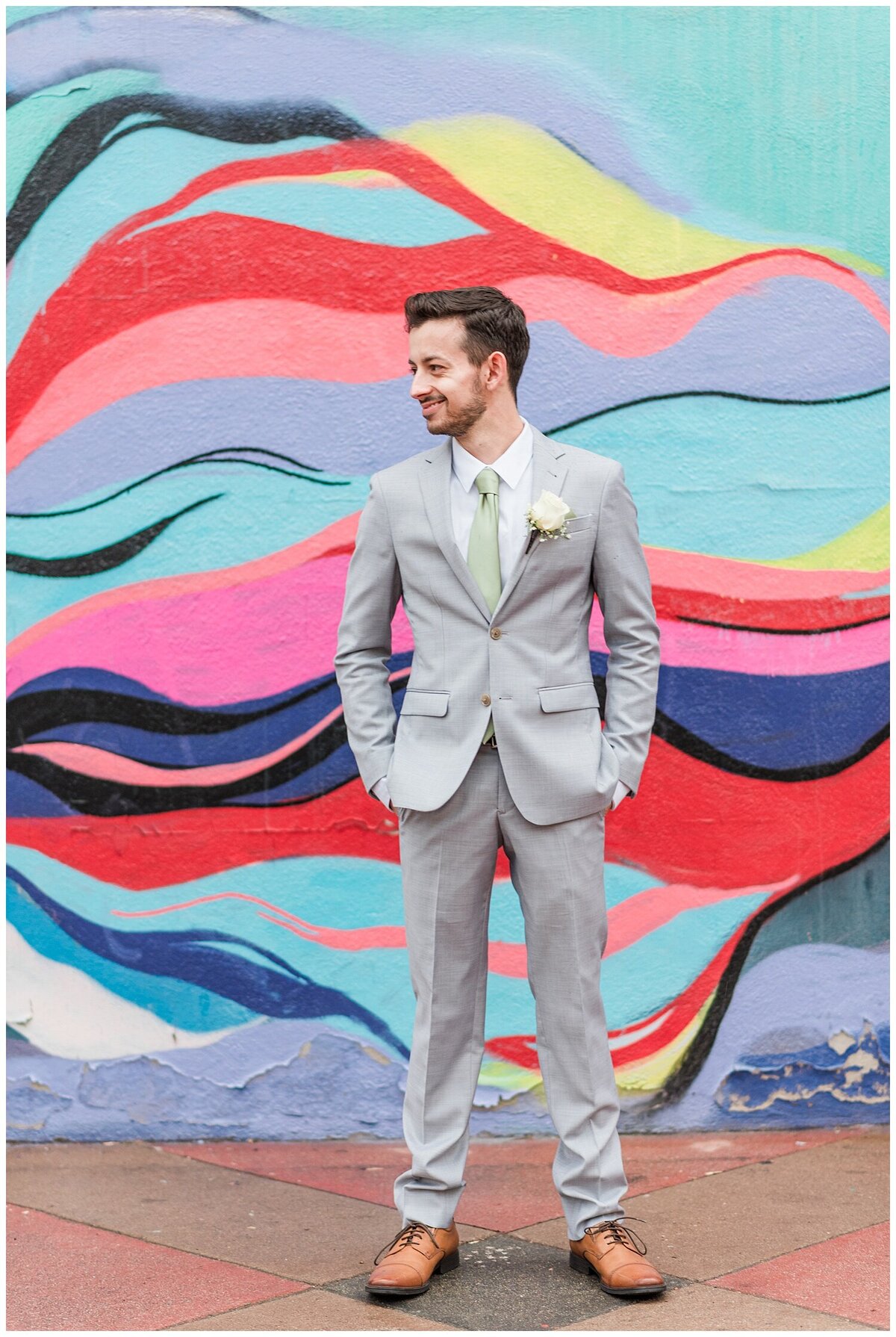 Groom in a gray suit with brown shoes stands square on with the camera and turns his head to the left with his hands in his pockets standing in front of a colorful wall in downtown Denver outside of Skylight venue