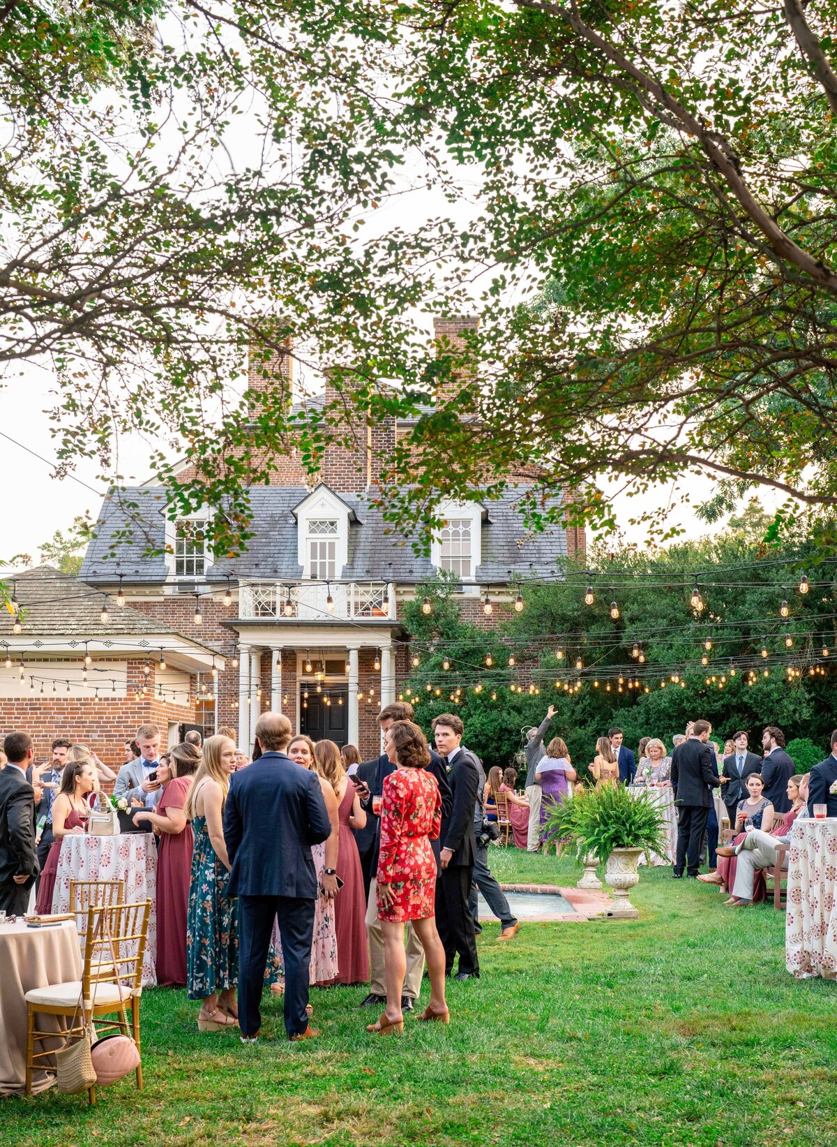 A lively outdoor garden party with people mingling under string lights. Guests are dressed in formal attire, gathered around tables with white tablecloths. A brick house with a porch is in the background, surrounded by greenery.