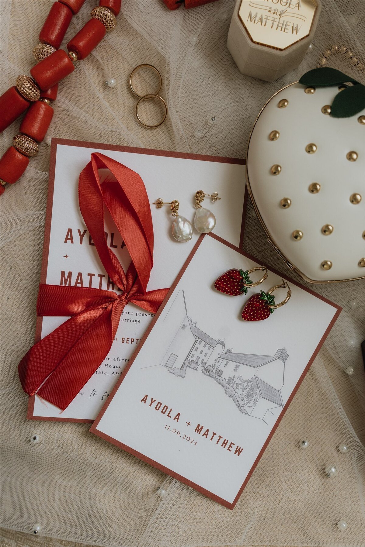 Wedding stationary is photographed together with wedding rings, jewellery and a brides veil. The stationary reads Ayoola and Matthew 11.09.24. Logie Country House is illustrated on the stationary.