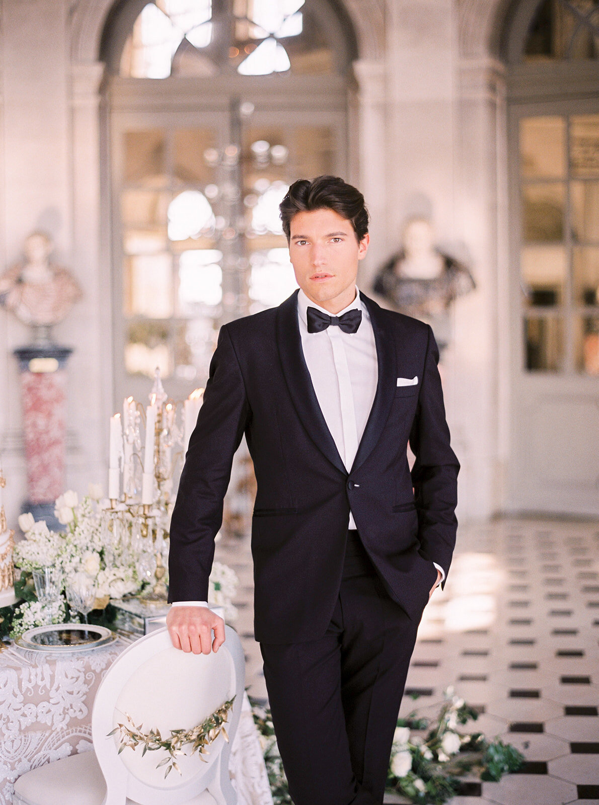 This stunning photograph was captured at the enchanting Chateau Vaux le Vicomte in Paris, France, renowned as one of the top destination wedding venues in the country. The talented photographer behind this exquisite shot is Audrey Fora. The image beautifully showcases a man in a stylish black tuxedo with a bow tie, standing gracefully in an elegantly decorated room. His hand rests casually on a chair adorned with delicate flowers, set amidst ornate architecture, tall windows, white floral arrangements, and a warmly candlelit table.
