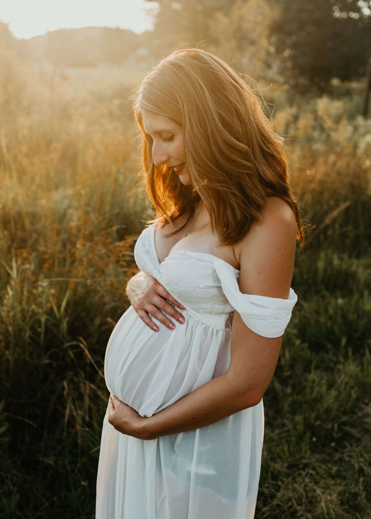 A pregnant woman in a white dress, captured by a Pittsburgh maternity photographer, standing in a field.