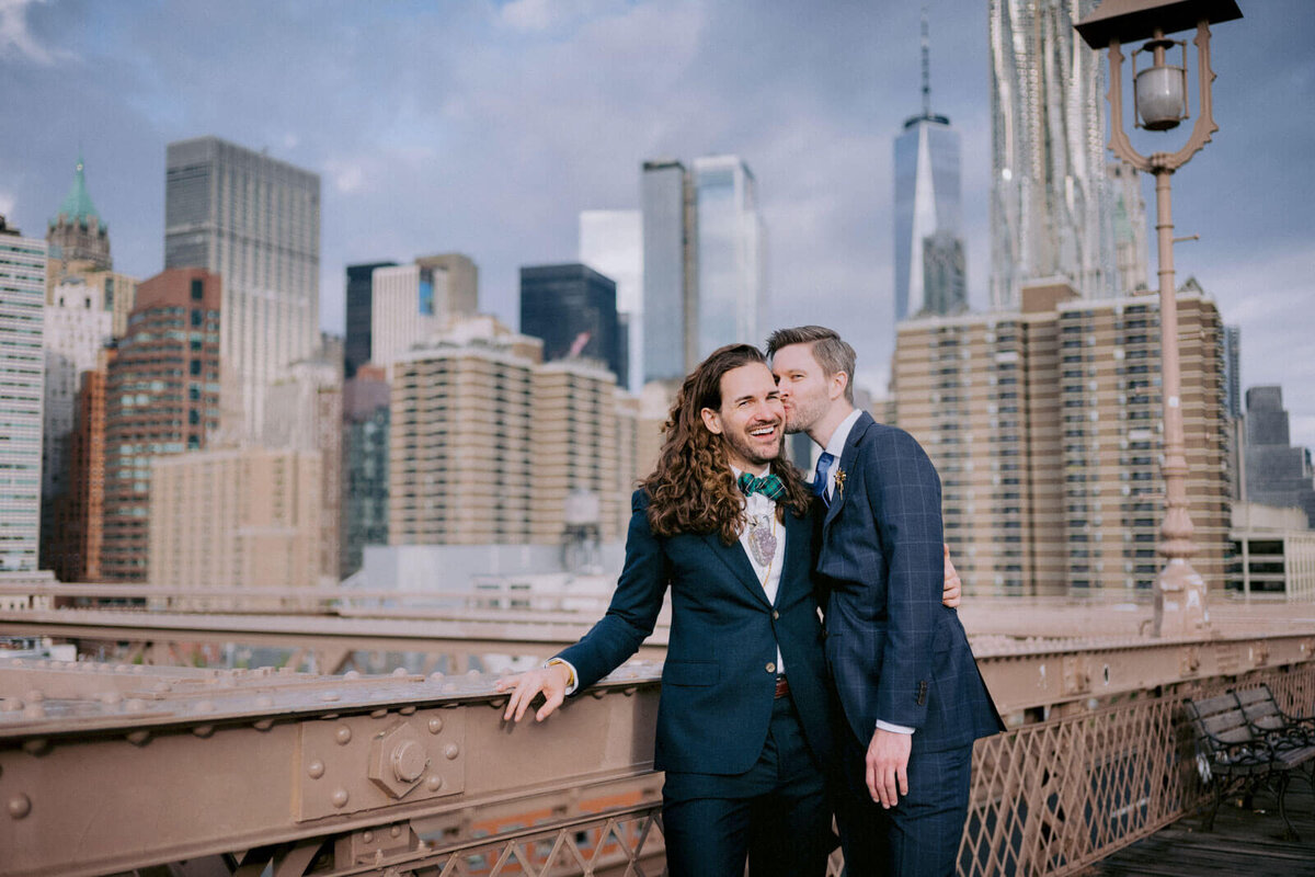 The groom is kissing his groom on the Brooklyn Bridge, with skyscrapers in the background. NYC Elopement Image by Jenny Fu Studio