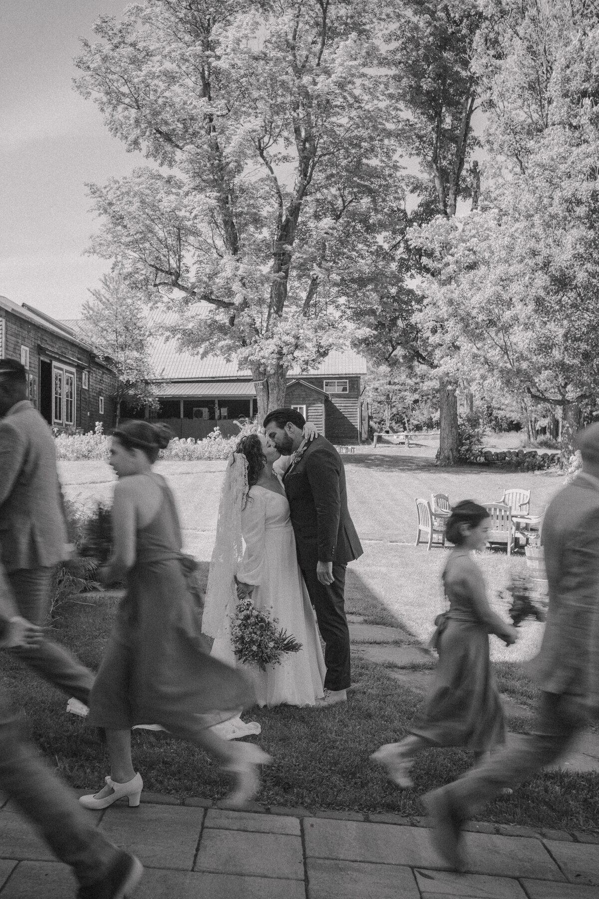 A black and white candid photo of a bride and groom sharing a kiss outdoors, with guests walking by in a blur. The serene setting features large trees and rustic buildings in the background, capturing a timeless and documentary-style wedding moment.