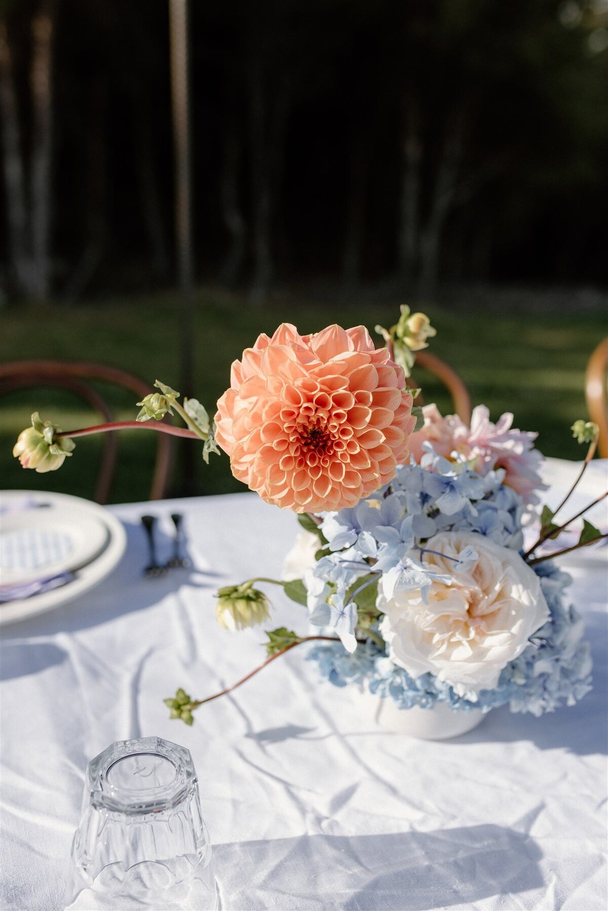 Bright orange dahlia centerpiece paired with pale blue hydrangeas and white roses on an alfresco reception table at a Lake Hāwea wedding.