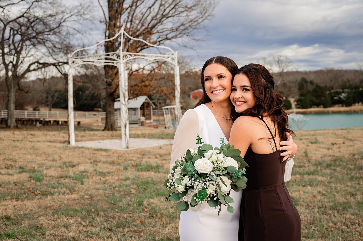 The bride, wearing a long satin wedding gown with long sheer sleeves holds a winter bouquet of ivory roses, white roses, seeded eucalyptus, blue eucalyptus, blue thistle, and silver dollar eucalyptus. She has one arm wrapped around her sister who hugs her back. The maid of honor is wearing a brown velvet dress with spaghetti dress. They are standing in front of a lake with a white metal gazebo at Steel Magnolia Barn.