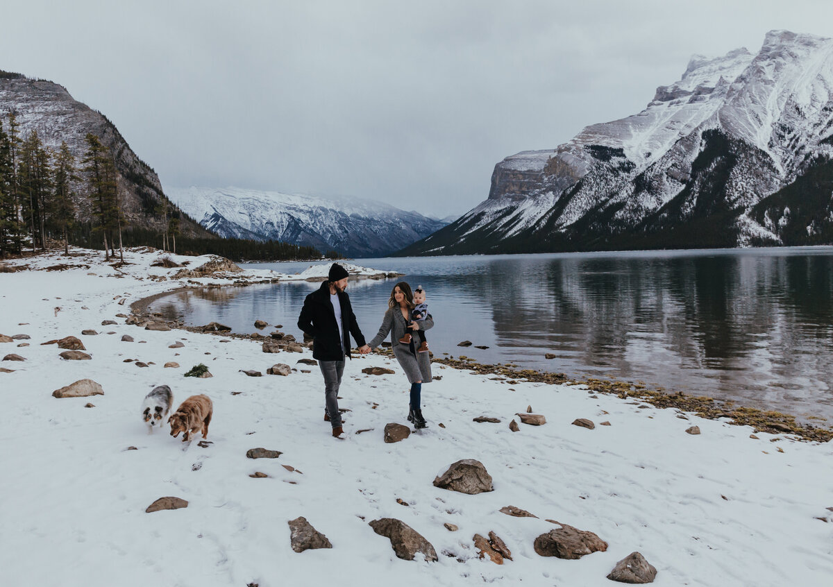 A mother and father walk hand in hand along a mountainside lake in the snow. The mother is holding her daughter on her hip and their dogs explore the lakeshore.