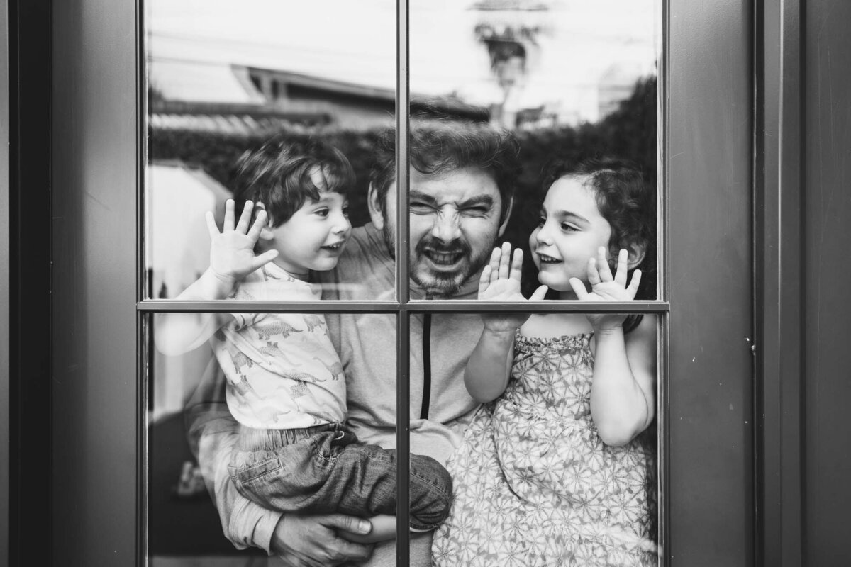Black & white shot through a window of a dad making silly faces while hi two children stare at him and smile.