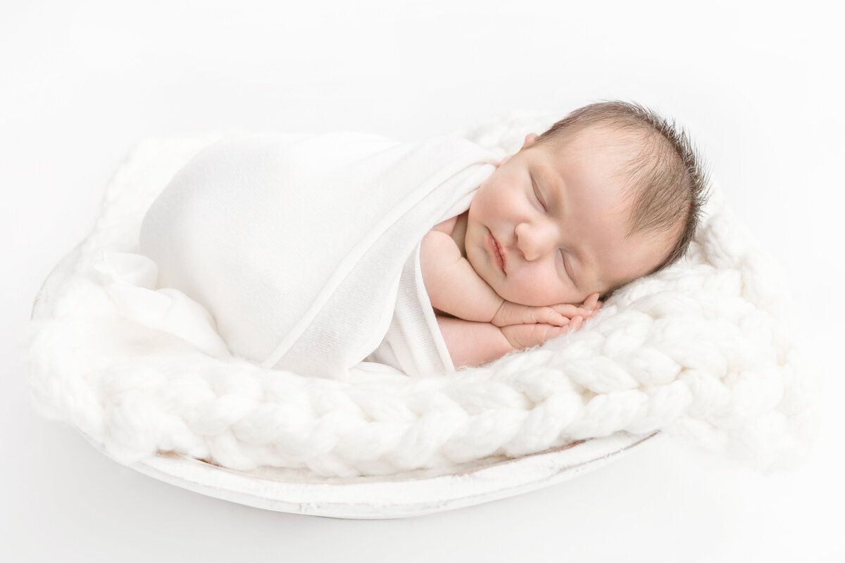 sleeping baby boy swaddled in white wrapped fabric lying on white fur inside a rustic handmade drift wood bowl for a posed studio newborn session