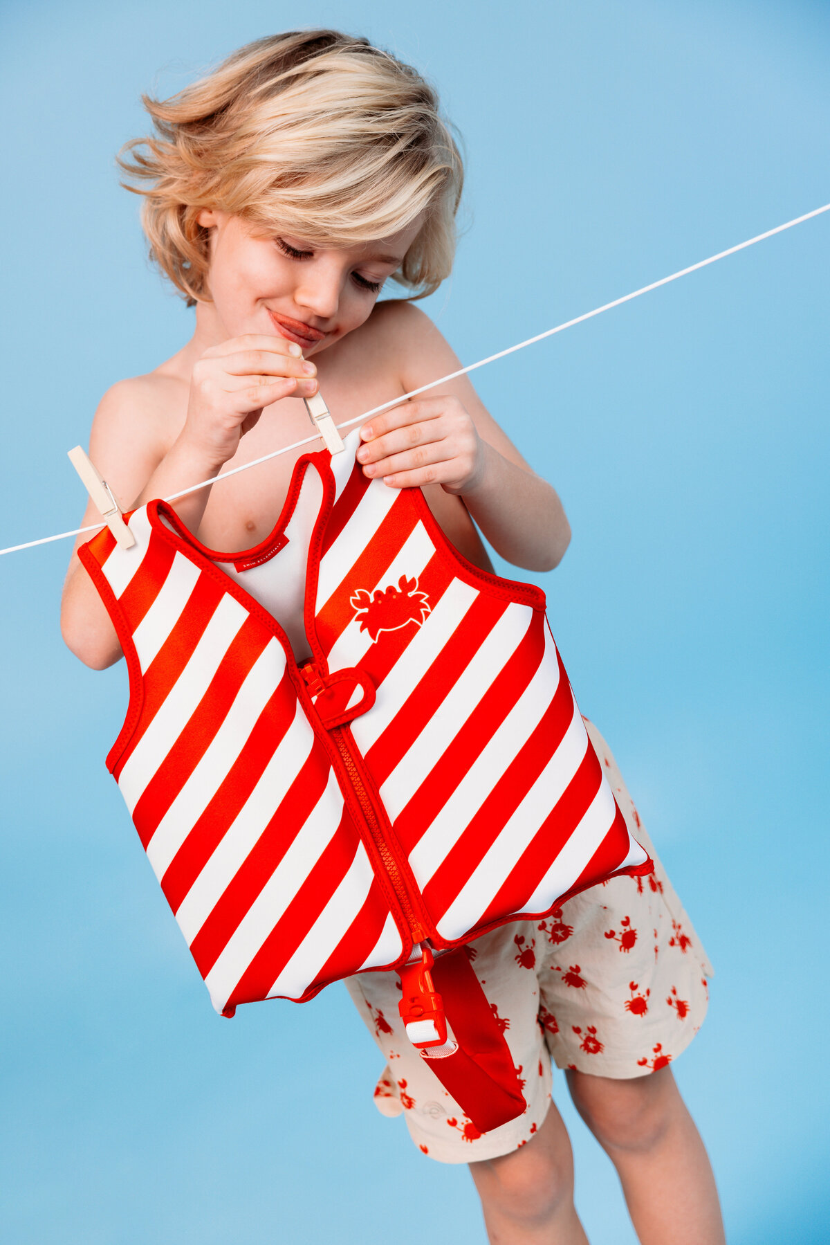 Smiling child holding a red-and-white striped Swim Essentials swim vest on a clothesline, wearing matching crab-printed swim shorts against a blue background.