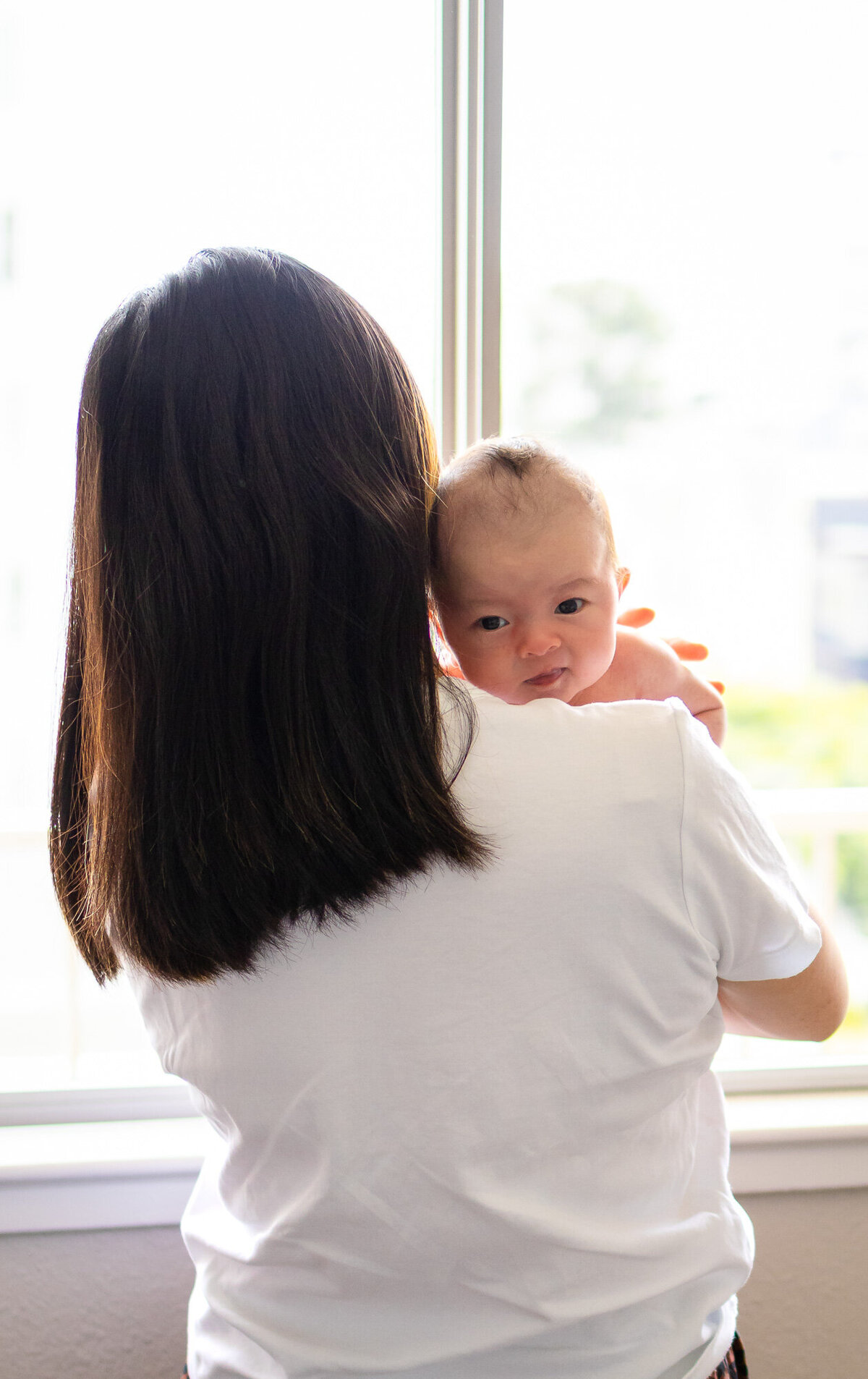 A newborn resting on mom’s shoulder, gazing out from the window light, captured by San Francisco newborn photographer.