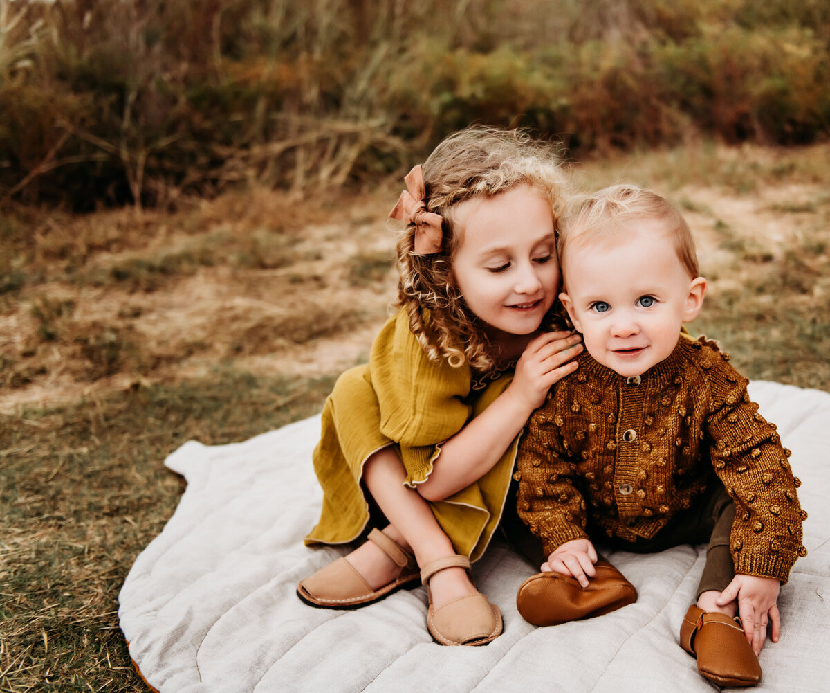 Little girl in a yellow dress snuggling her baby brother on a blanket.