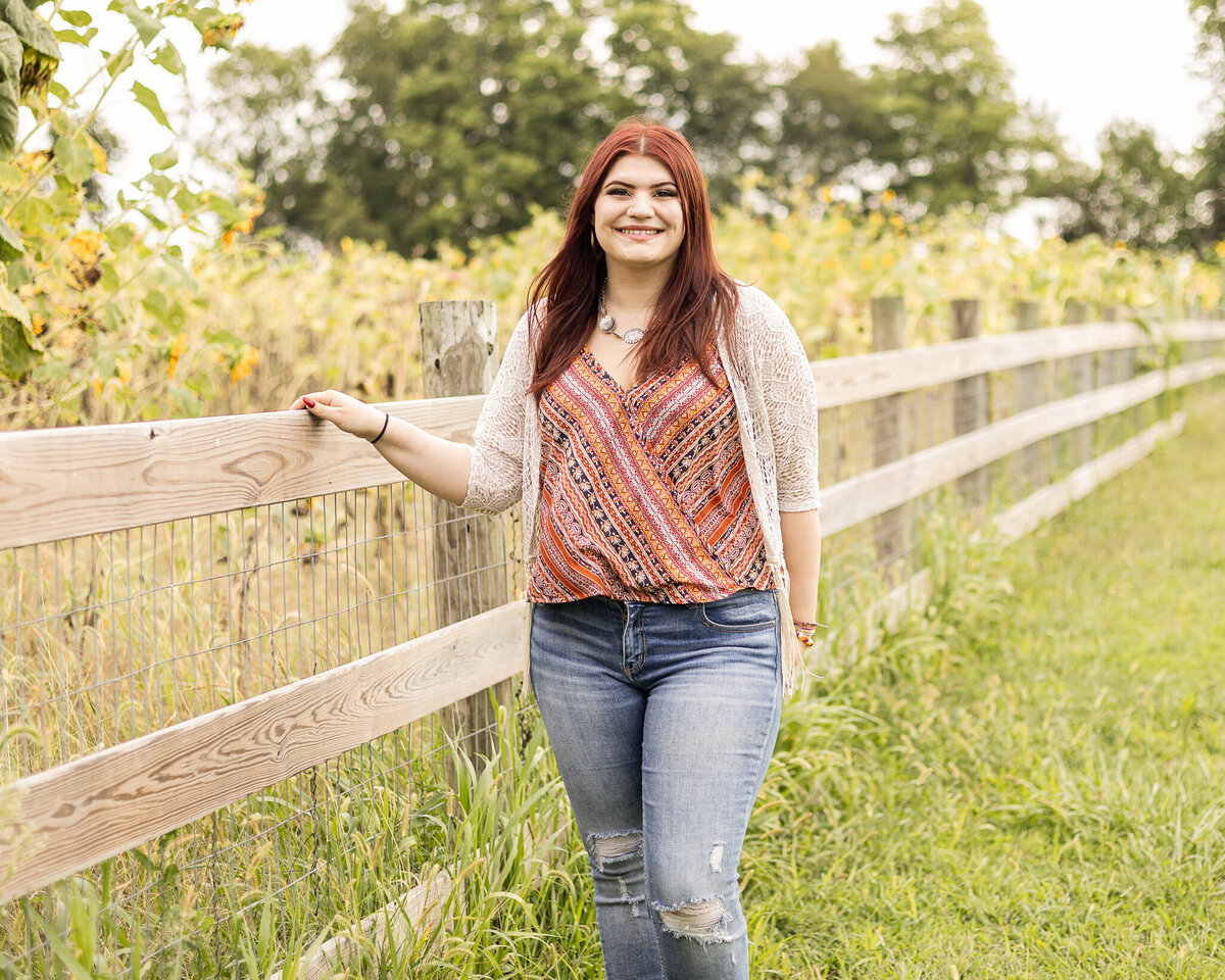 High school senior female with long red hair, wearing blue jeans, light tan smock over a patterned shirt, standing in the grass, next to a fence with flowers in the background smiling to the camera
