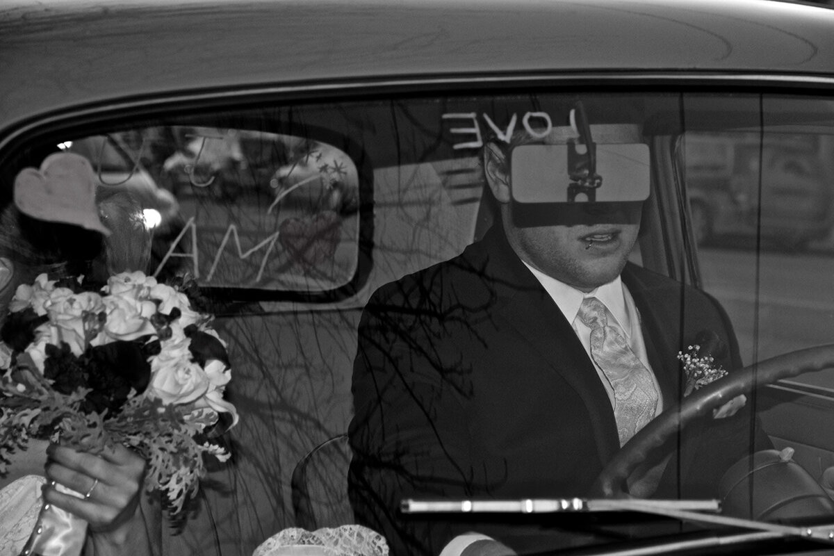 Black and white portrait Justin and Katie Wilkie front seat of car through windshield rear view mirror covering grooms eyes on their wedding day Reflections Gallery