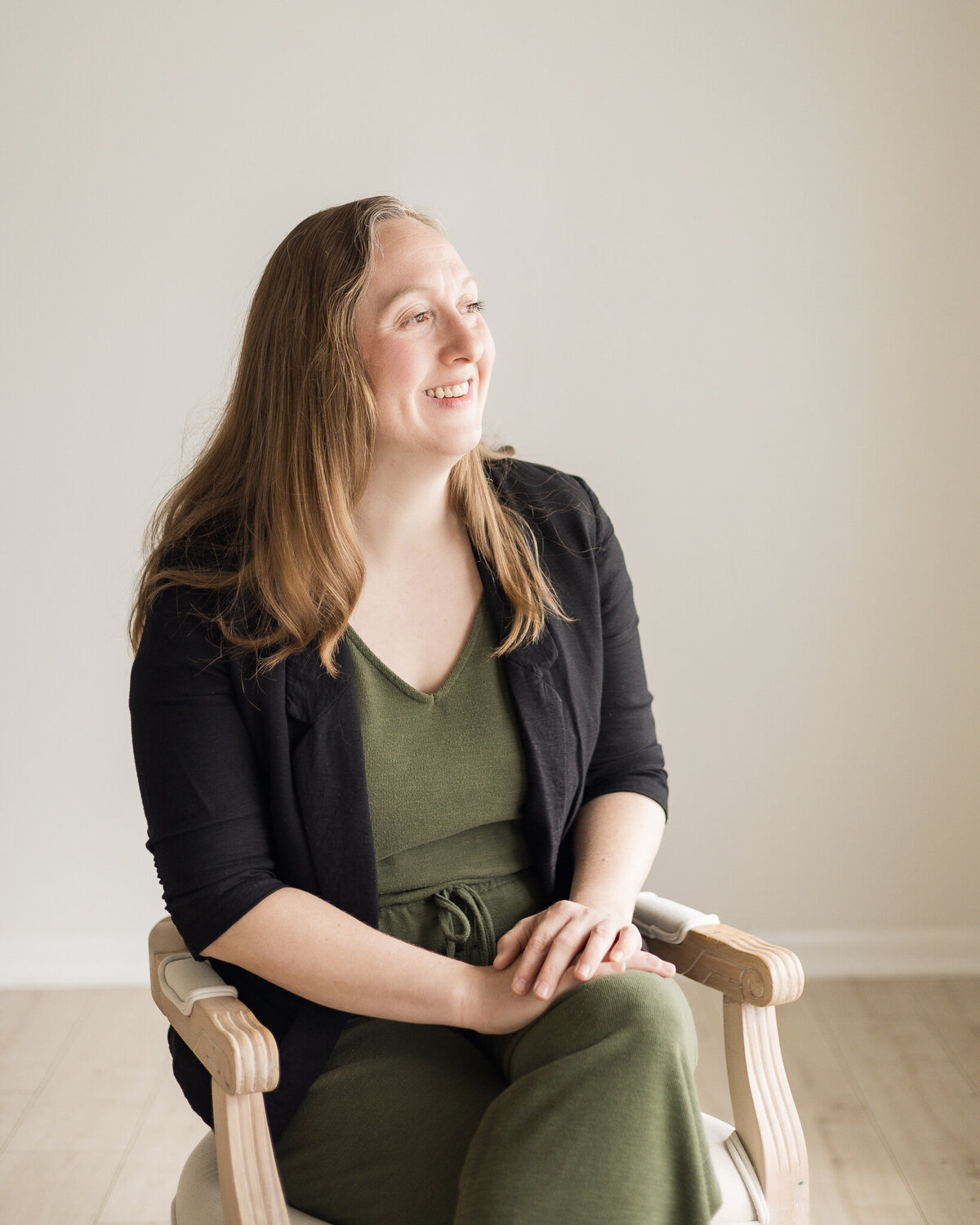 Strawberry blonde female, sitting in a chair, wearing dark green pants and shirt with a black jacket over the green outfit, smiling off to the side of the camera