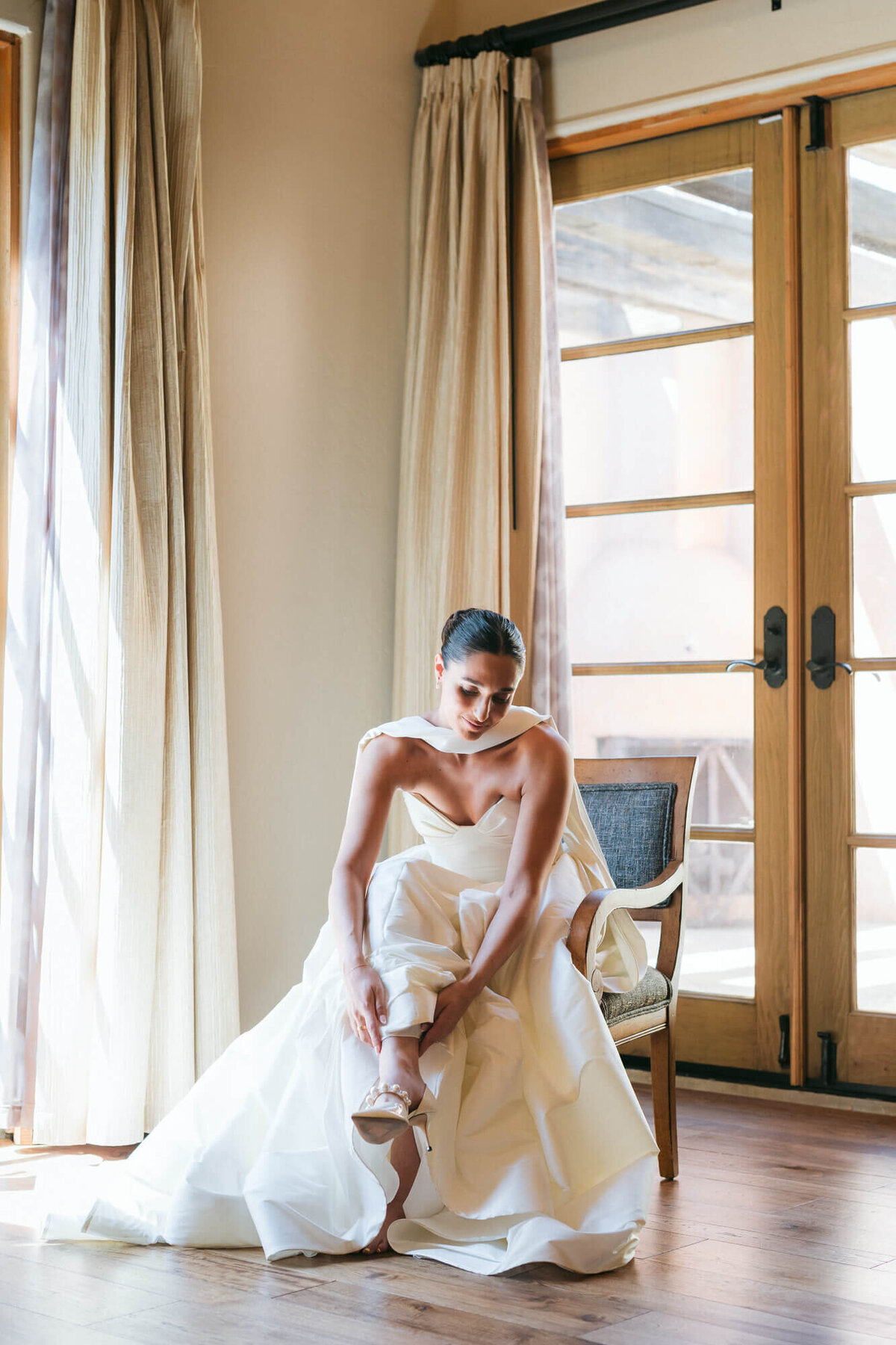Bride in a strapless gown putting on her heels near soft morning light before her Phoenix wedding, captured by a Phoenix wedding photographer.