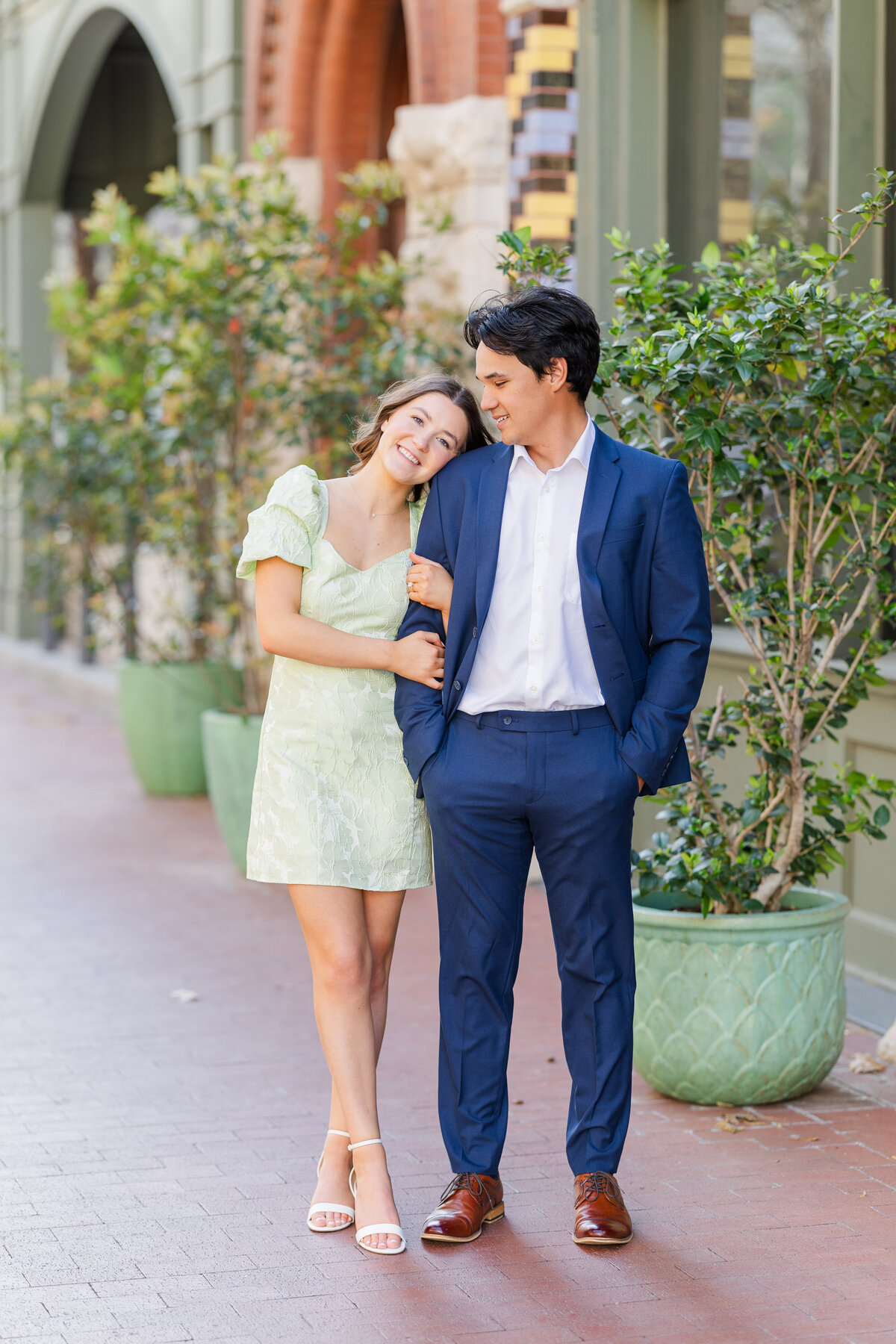 A fashionable couple poses during their downtown Fort Worth engagement shoot, with the bride-to-be resting her head on his shoulder. She wears a light green floral dress while he’s dressed in a sharp navy suit, creating a classic and elegant city vibe. A romantic moment captured among the charm of Fort Worth architecture.