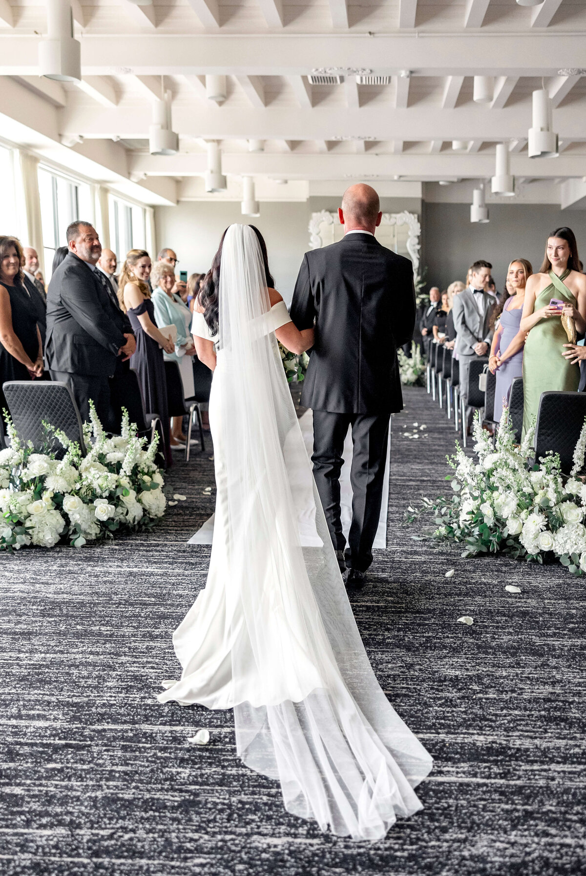 A bride in a flowing white dress and veil walks down the aisle with a man in a black suit. Guests are seated on either side of the aisle lined with white flowers, watching the procession in a bright, modern venue.