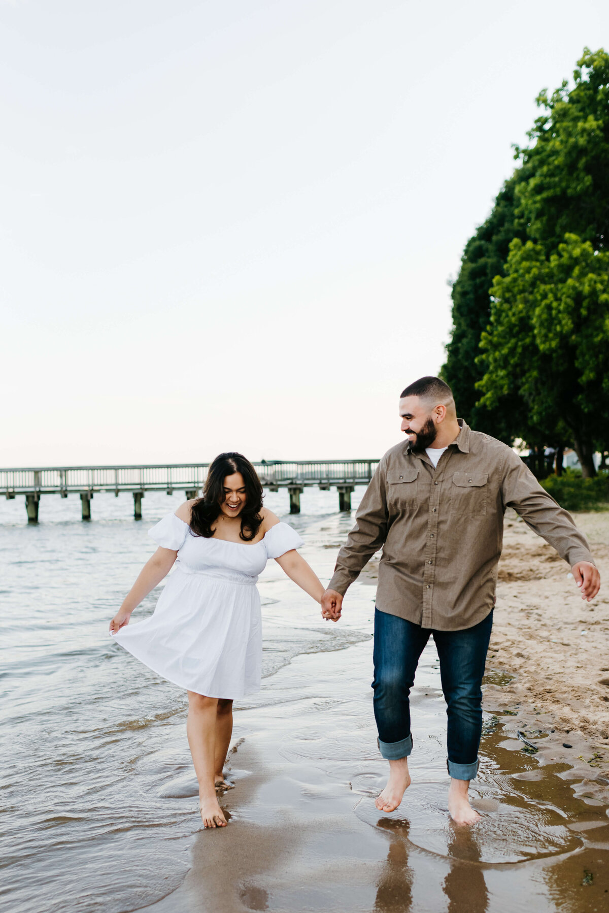 romantic engagement photos with man and woman holdin ghands while walking in the water at  woodbridge lake beach with a bridge in the distance