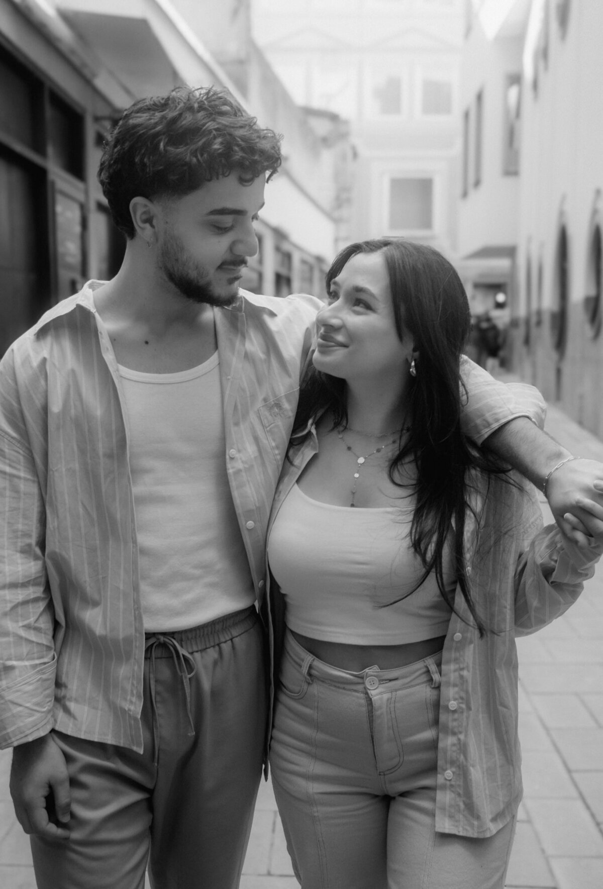 The couple walk down a street in a casual embrace and look into each other's eyes, visibly in love.