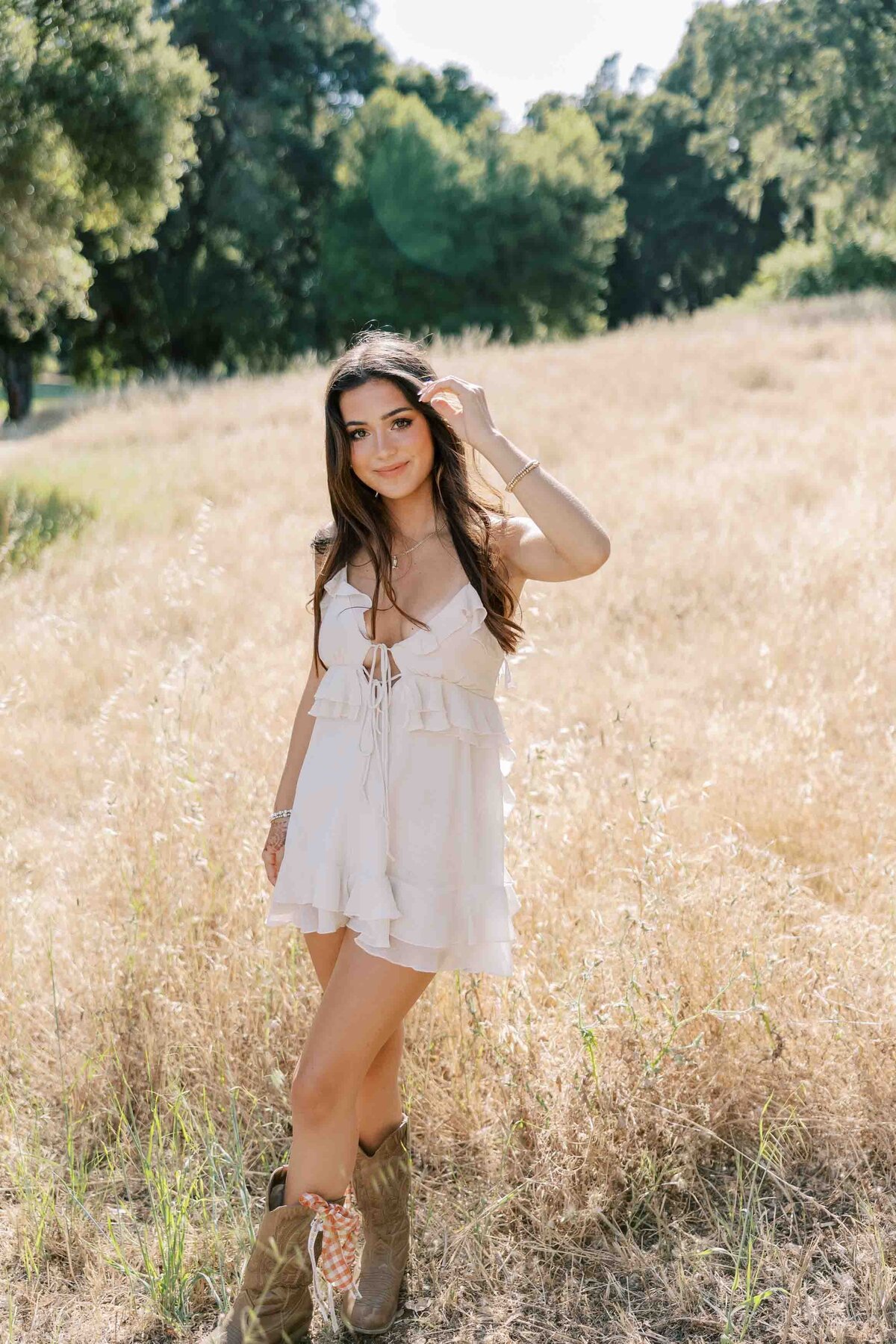 Brunette girl smiling with a soft smile at the camera, wearing a white dress and standing in an area with dry grass