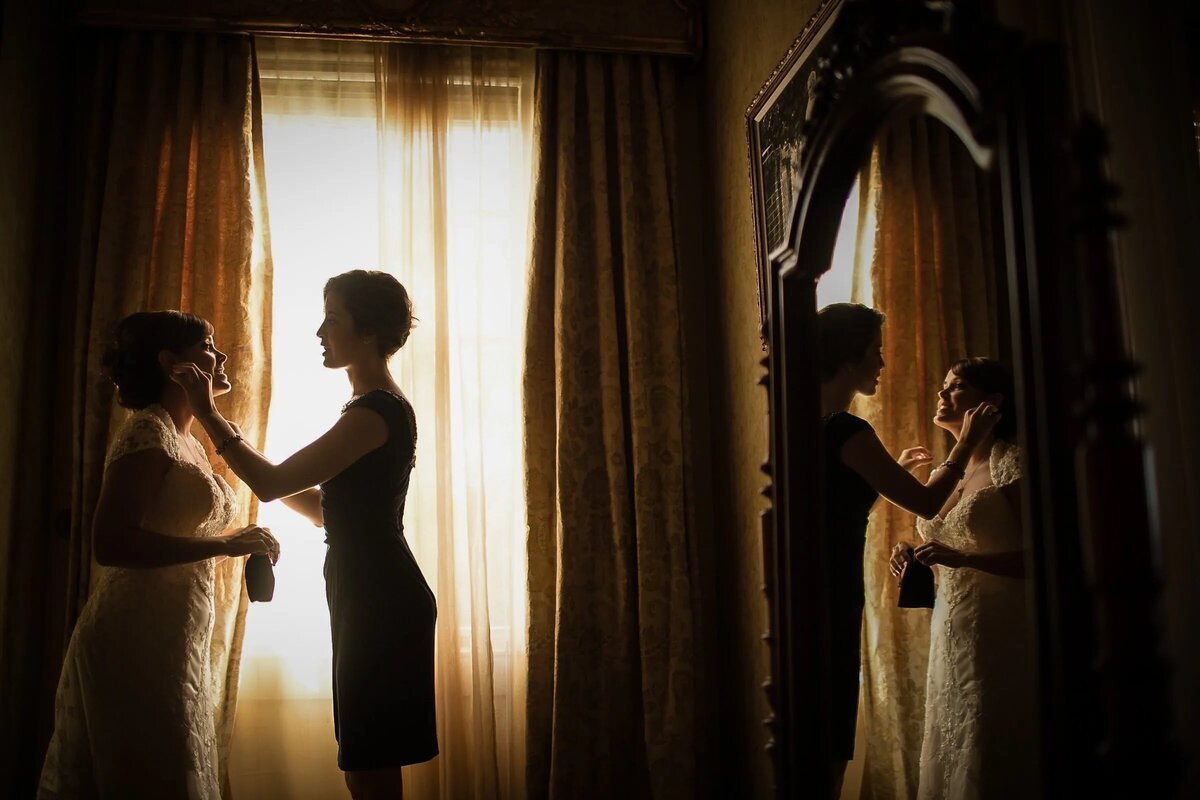Warmly lit image of a bride being assisted by a bridesmaid, with a mirror reflection