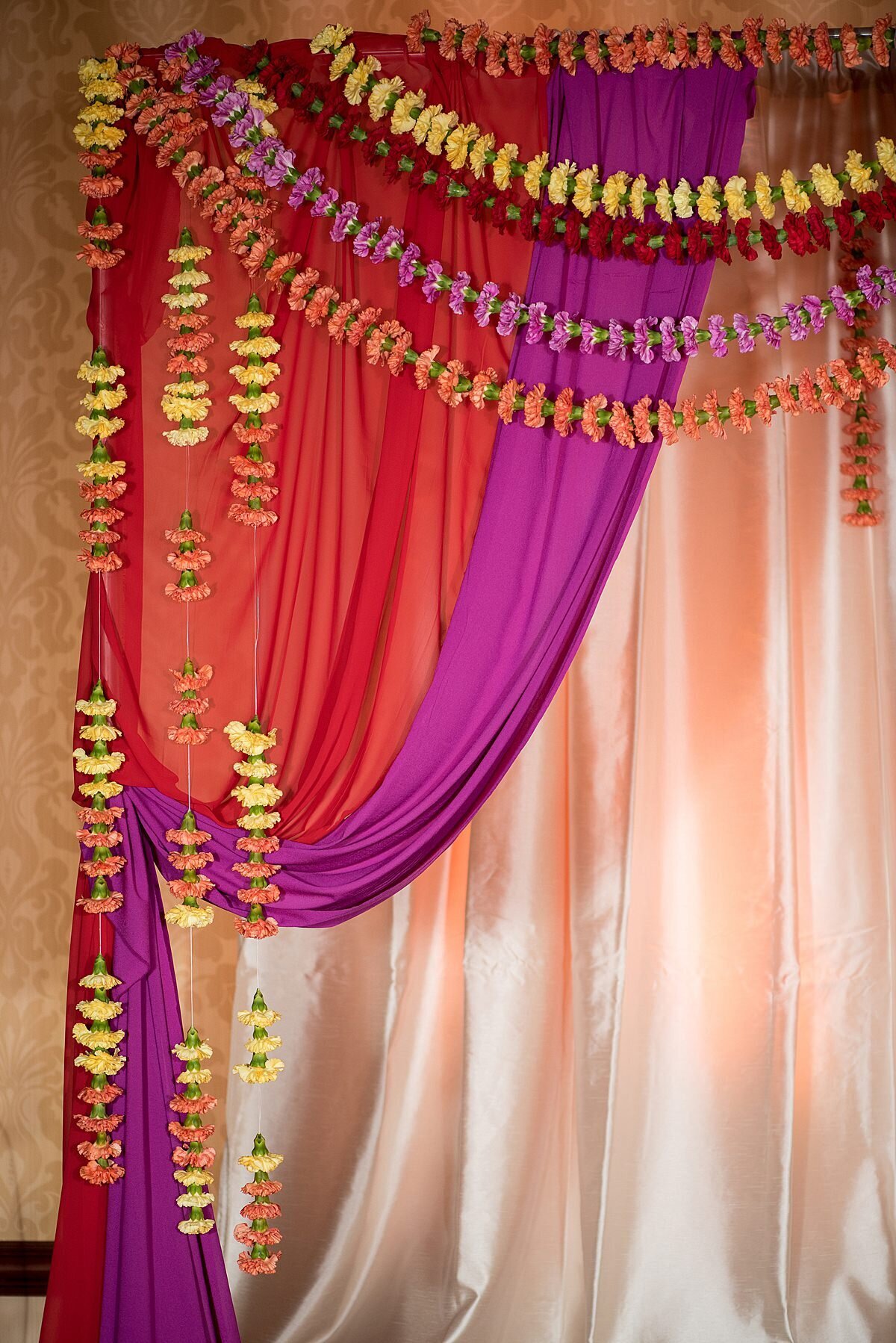 Strands of orange and yellow carnations hanging on the edge of a red and purple mandap with ivory drapery.
