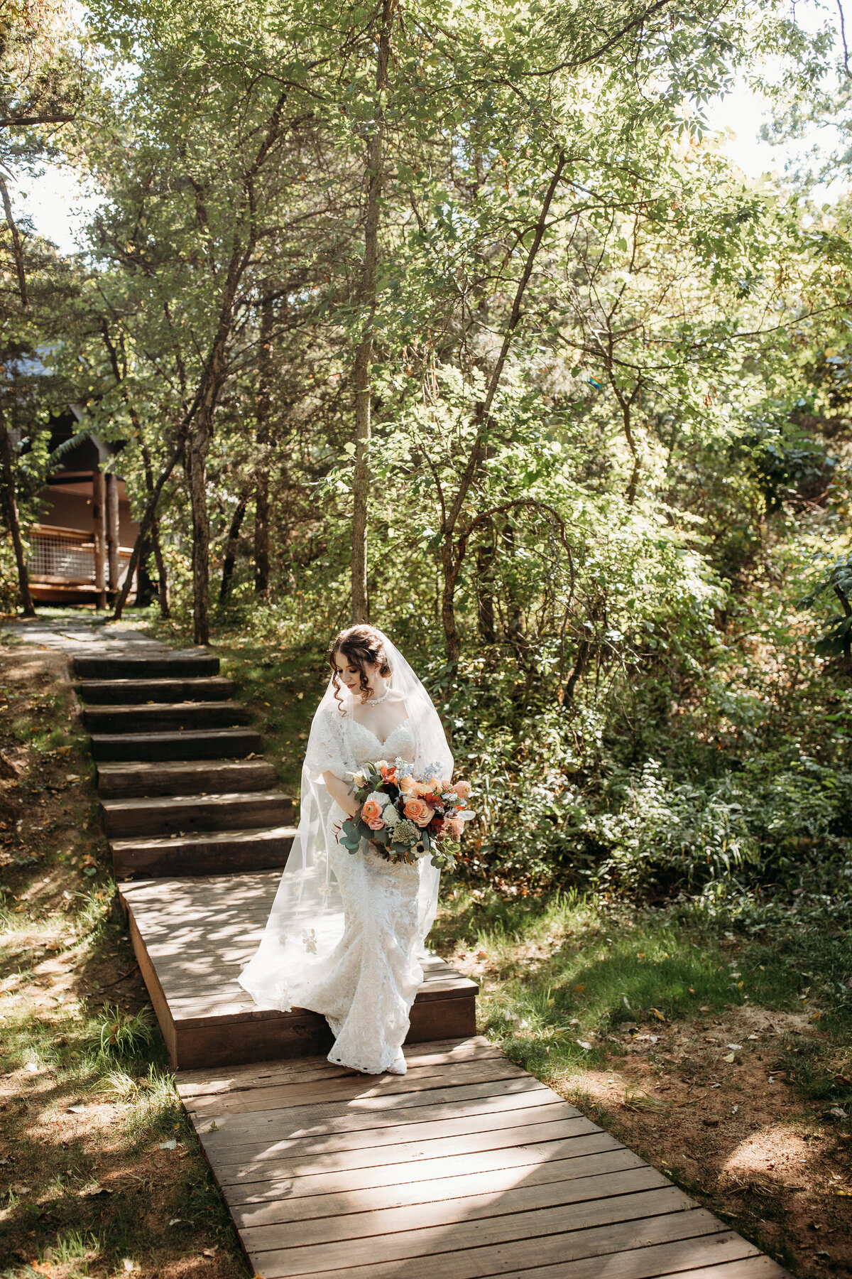Bride descending a wooden stairway in a forest, sunlight streaming through the trees