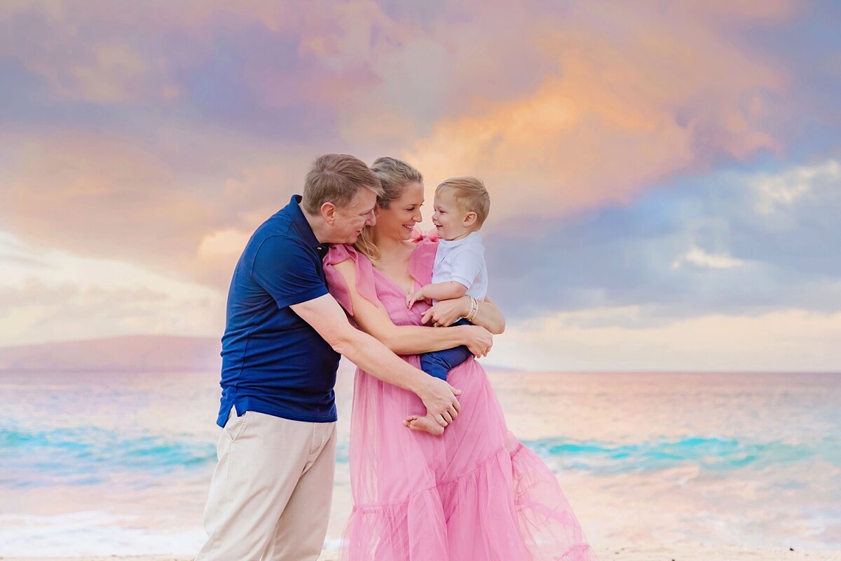 Mother in pink dress and dad in blue button down shirt hold their son and look at him during a sunrise family session in Maui