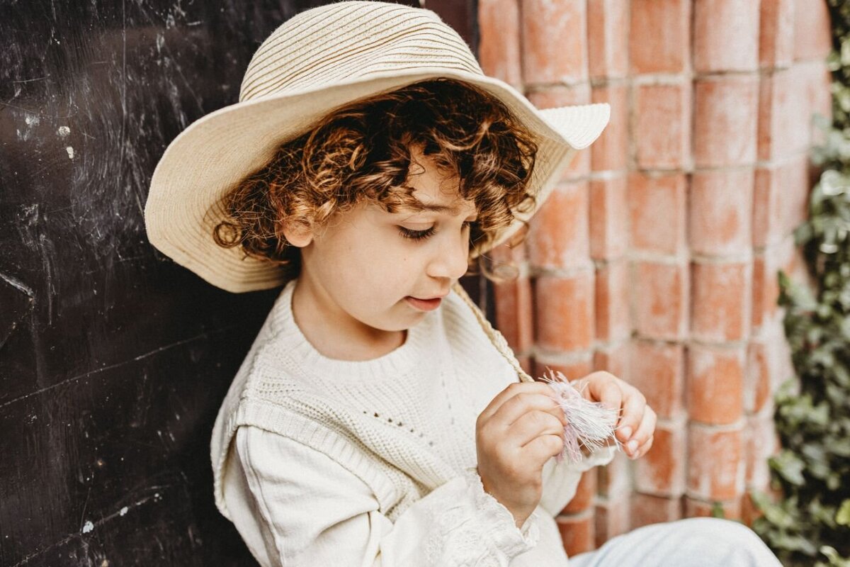 A young child with curly hair wearing a striped sun hat thoughtfully examines a small object in their hands while sitting against a textured wall 