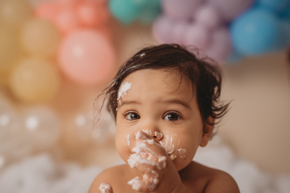 Photo shoot at Atlanta, Georgia cake smash photographer studio with one year old little girl in pink tutu smashing a cake with rainbow pastel balloon garland behind her with white fluffy clouds.
