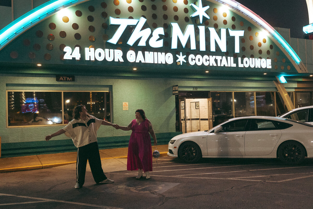 A couple playfully reaching out to each other under the neon lights of 'The Mint' casino sign