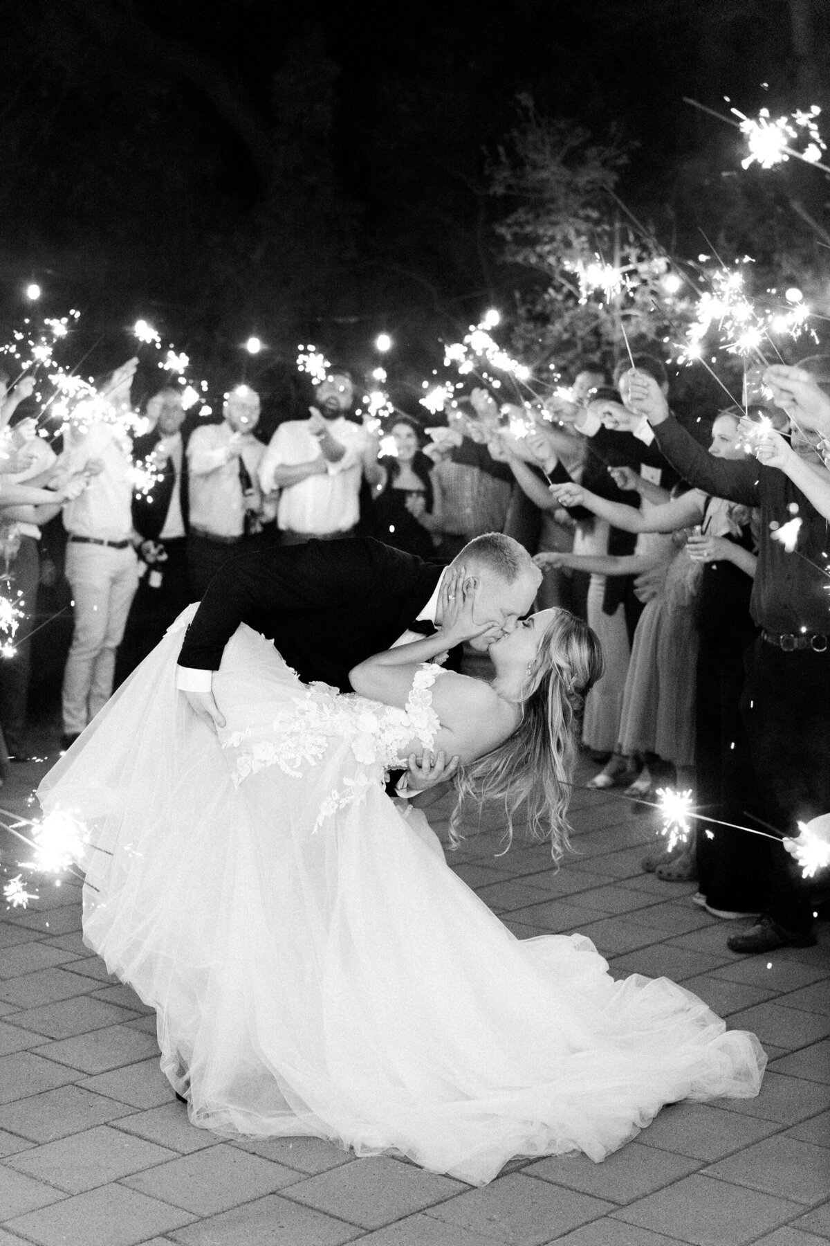 A couple dressed in wedding attire shares a kiss while the groom dips the bride. Guests surround them holding sparklers, creating a celebratory atmosphere that highlights their wedding in Canada.