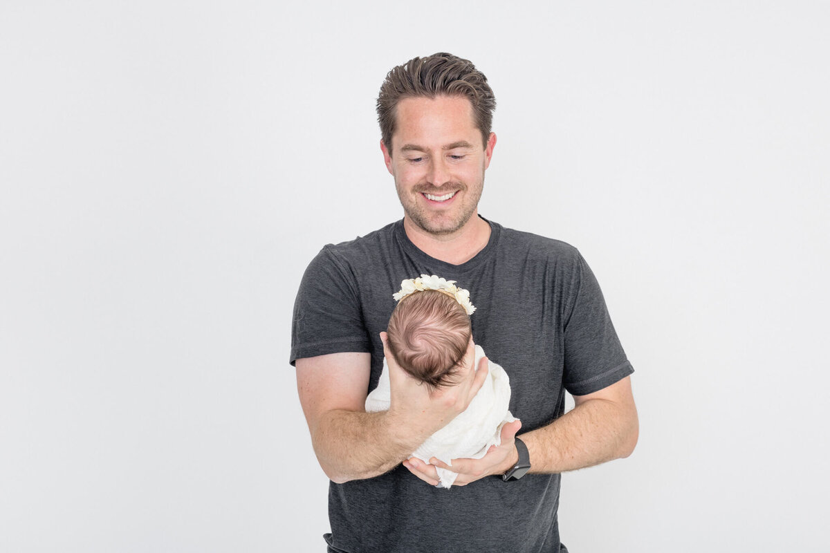 A man with short hair, wearing a dark t-shirt, smiles while holding a baby wrapped in a white blanket. The background is plain white.