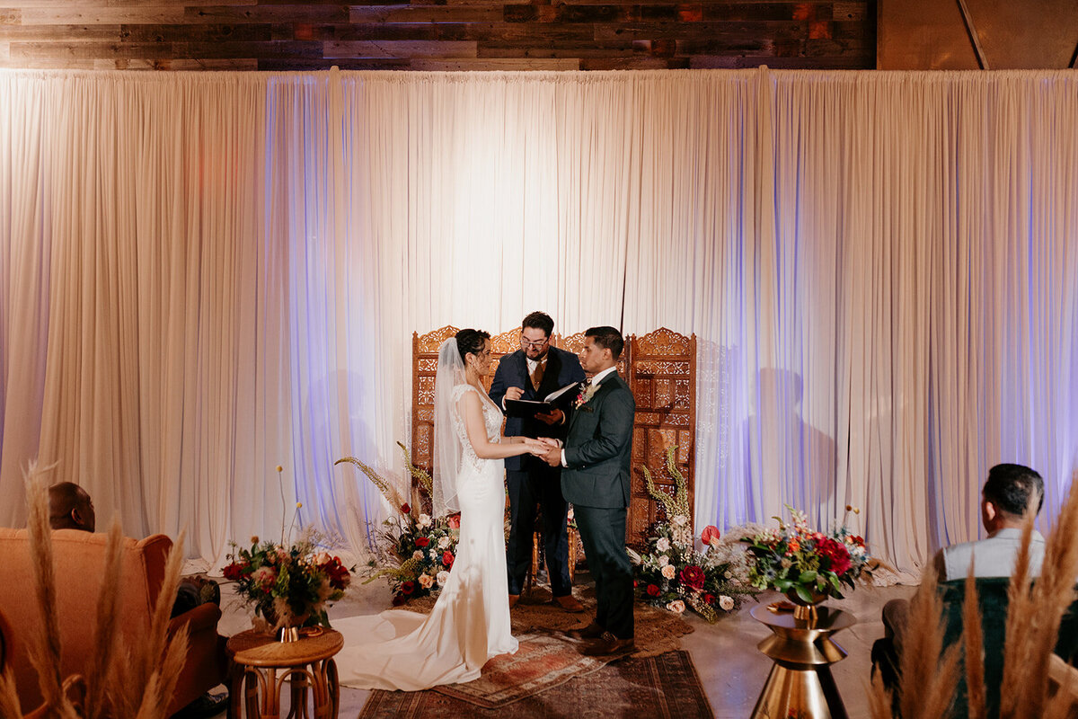 Wide shot of the wedding ceremony, showcasing the couple holding hands in front of an officiant with soft drapery and floral decorations enhancing the setting.