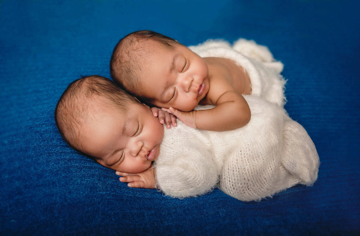 Newborn photos of the Greater Toronto, ON area, twins in white sleeper cuddling together on a blue backdrop.