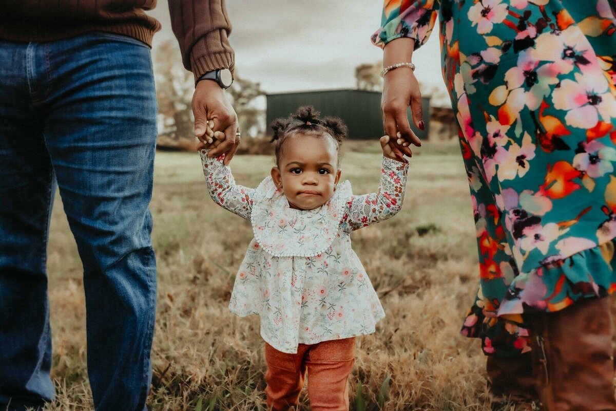 A toddler walks on grass, holding hands with two adults. The child wears a floral top and orange pants. The adults' clothing is partially visible.