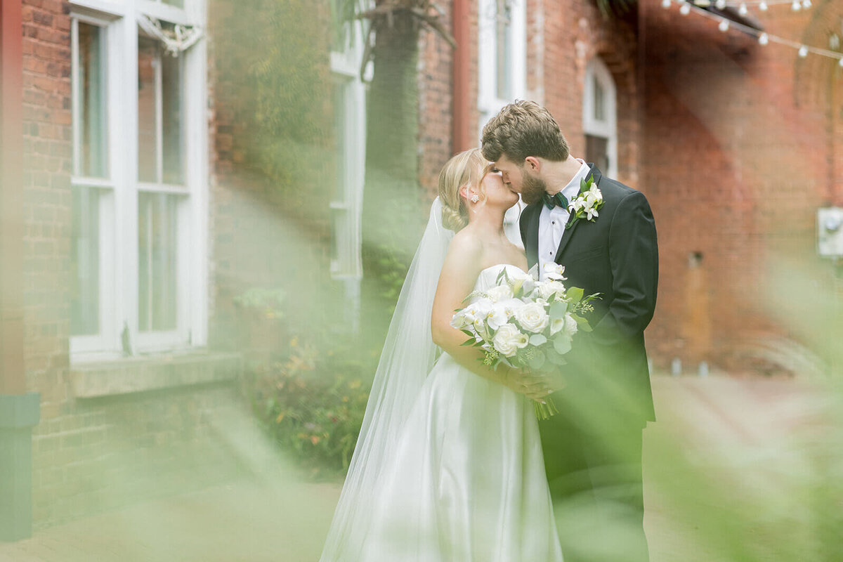 A romantic wedding portrait at 701 Whaley captures a newlywed couple sharing a kiss, framed by soft, blurred greenery. The historic brick architecture and string lights in the background add warmth and charm, while the bride’s elegant satin gown and classic bouquet complement the groom’s timeless black tuxedo. The natural light and dreamy foreground create a beautifully intimate and timeless image.